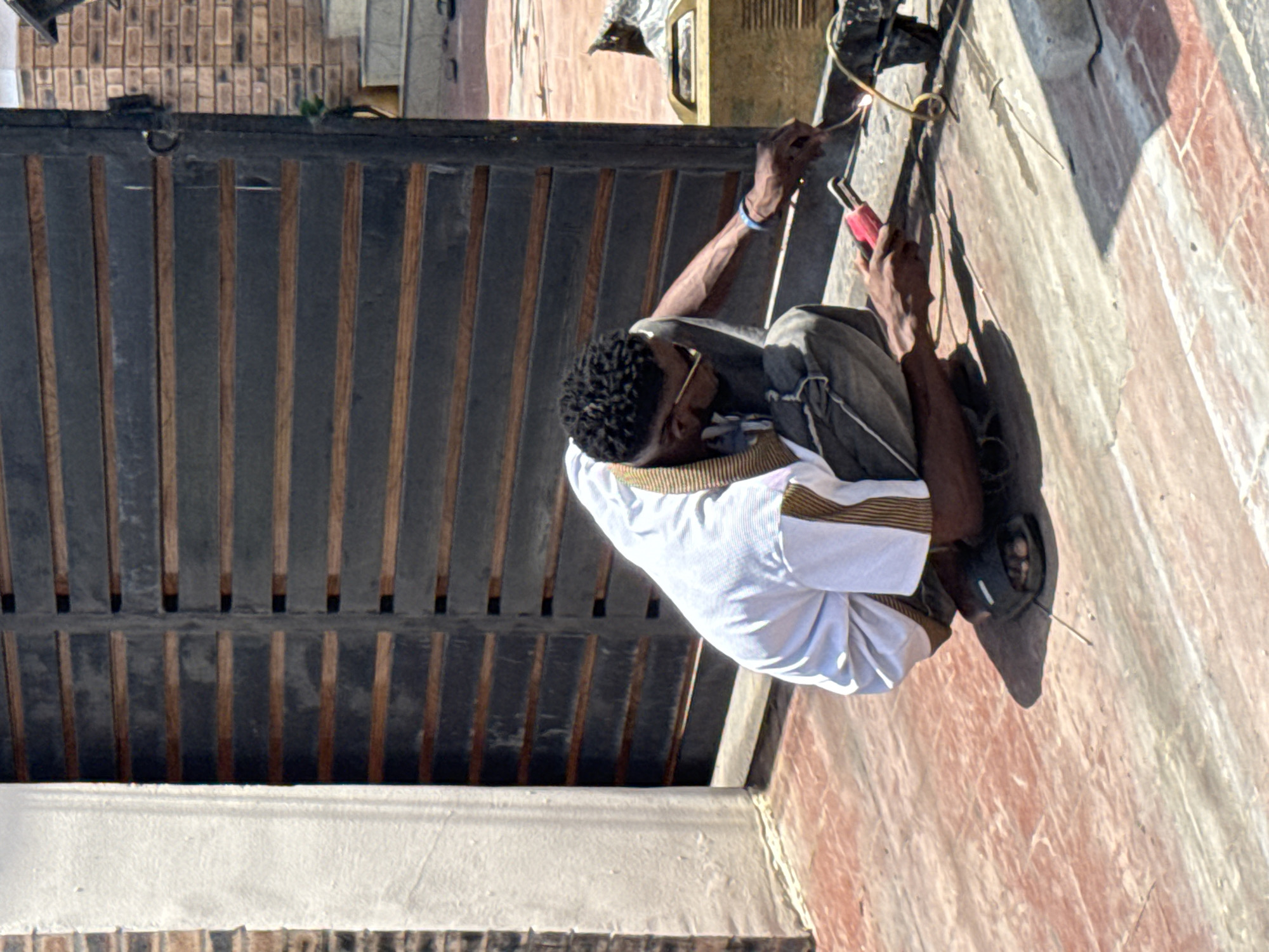 A man wearing a white shirt, dark pants, and sandals is squatting and working on a black metal gate. He is using a tool with a red handle to work with wires near the top of the gate. The gate is made of vertical slats, and a building with brickwork is visible in the background. The scene appears to be set on a concrete or stone surface, possibly a sidewalk or driveway. The location is in Lekki, Nigeria.