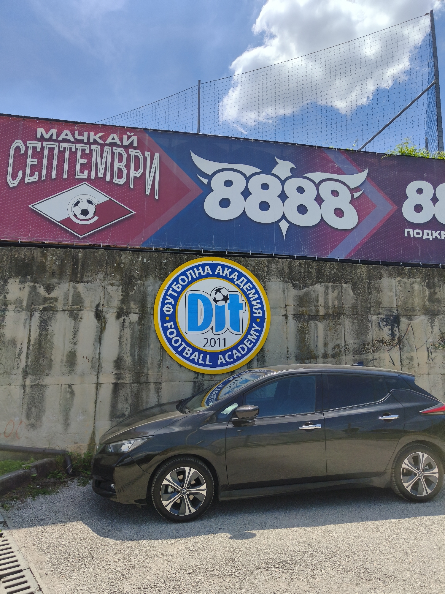 The image shows an outdoor scene on a sunny day with a blue sky and scattered clouds. In the foreground, a dark-colored electric car, possibly a Nissan Leaf, is parked on a gravel surface. The car is facing towards the right of the frame. Behind the car, there is a concrete wall. Mounted on the wall is a circular logo with a yellow border, containing blue and white elements. The logo features text in a circular arrangement, with "DIT" prominently displayed in the center, along with the year "2011" below it. Above the concrete wall, a large banner is visible, displaying text in Cyrillic and Arabic numerals. The text "МАЧКАЙ СЕПТЕМВРИ" is prominently displayed on the left side of the banner, above a logo that appears to be a diamond shape containing a soccer ball. The right side of the banner features large white numbers "8888". The banner is stretched across a fence. The environment appears to be a sports facility, likely a football (soccer) stadium or training ground, given the presence of the soccer-related imagery and the fence. The overall impression is a candid shot capturing a car parked in front of a facility with prominent branding. This location is the DIT Football Academy, situated in Sofia, Bulgaria, specifically near the Vasil Levski National Stadium.