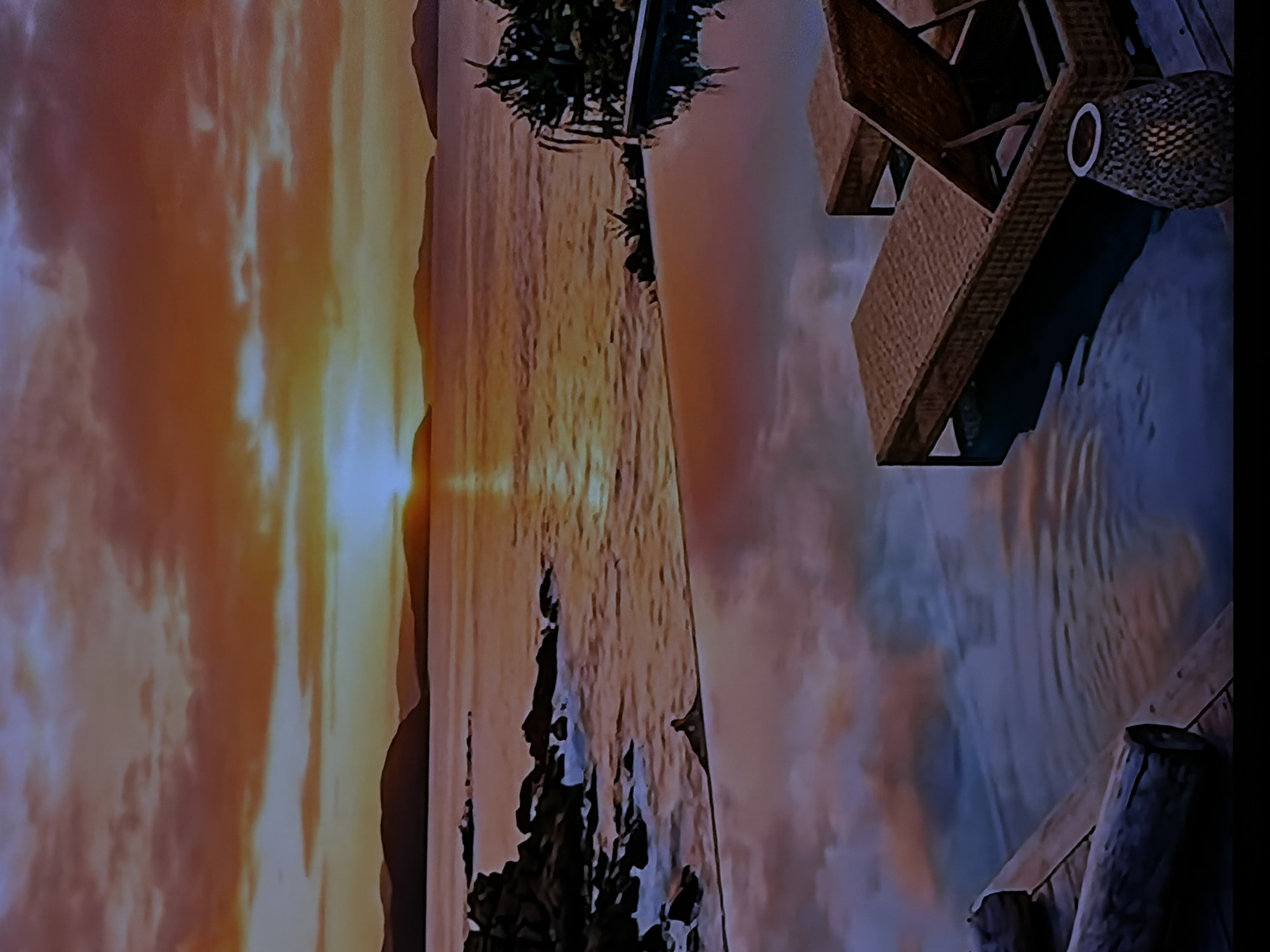 The image displays a serene landscape at what appears to be either sunrise or sunset, viewed from a structure extending over water. The scene is dominated by a vibrant sky, bursting with warm hues of orange, yellow, and red from the low-lying sun, which casts a bright glow on the horizon and reflects as a shimmering path on the water's surface. Dramatic clouds are scattered across the sky, transitioning to cooler blue and purple tones.
In the foreground, on the right side of the image, two lounge chairs or sunbeds, constructed from what appears to be woven wood or wicker, are positioned on a deck or pier that overlooks the water. The chair closest to the viewer features noticeable textural details and includes a cylindrical accessory with a white ring. Wooden posts or supports for the structure are visible beneath the deck. On the far side of the calm body of water, dark silhouettes of trees or dense vegetation line the distant shore, with one tree being more distinct in the middle-left of the frame. No individuals are present in the scene. The overall impression is one of tranquility and relaxation. There is no visible text.
Given the location context of Sofiivska Borshchahivka, Ukraine, it is highly probable that this image is a photograph of a screen or projection displaying a scenic vista, rather than a direct outdoor view from that geographical location.