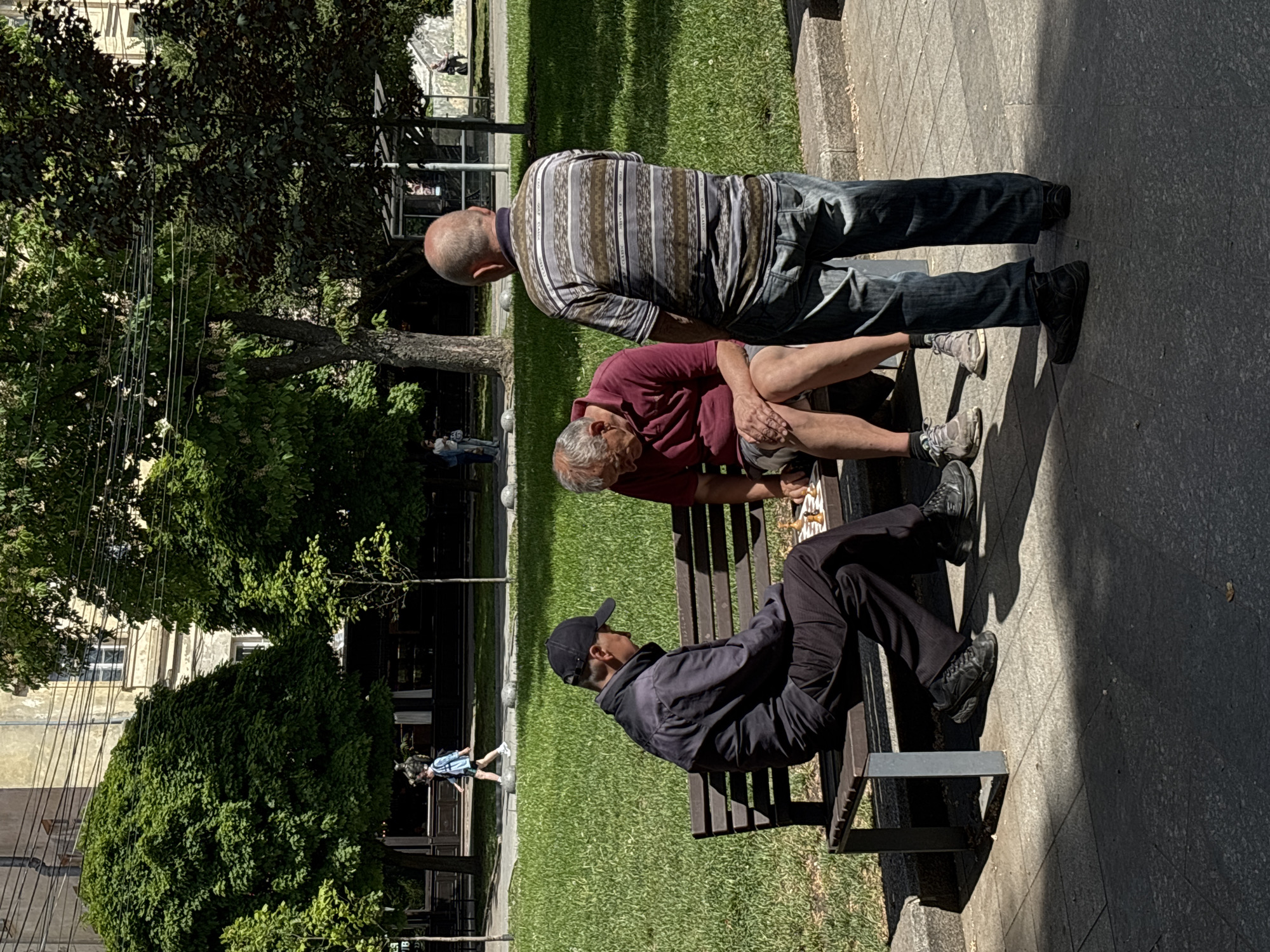 The photograph shows three men in a park, likely in Lviv, Ukraine. One man is sitting on a bench, wearing a black cap and jacket and dark pants. Another man is sitting on the bench beside him wearing a maroon shirt and shorts. The third man is standing behind the two, wearing a horizontally striped shirt and jeans. It appears they may be playing chess. The bench sits on a cement walkway, next to a grassy area. Trees and a building are visible in the background. A person walks near the building on the left.