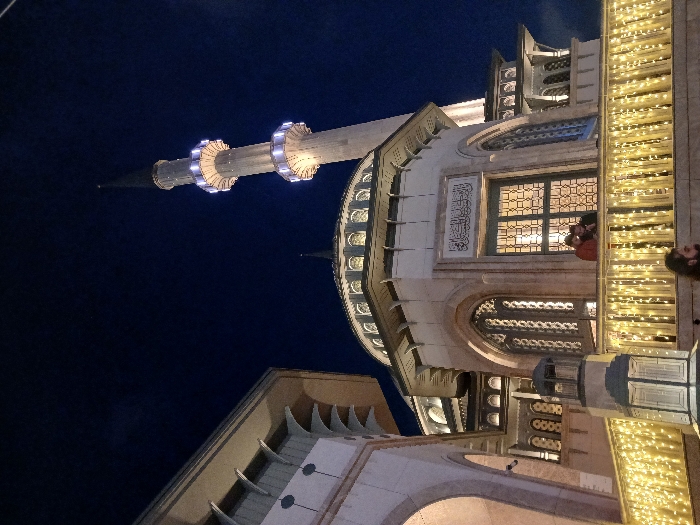 A nighttime view of the exterior of the Eyüp Sultan Mosque in Istanbul, Türkiye (latitude: 41.037, longitude: 28.984). Two men stand on a balcony facing the viewer, and the balcony is decorated with white string lights. The mosque is lit up, the minaret is tall and narrow, and the sky is dark. The mosque is lit up, and the minaret is tall and narrow. The sky is dark, and the only lights visible are the streetlights. There is a large window in the center of the mosque, and intricate detailing on the building. The mosque is located in the Eyüp district of Istanbul. It is a popular destination for Muslim pilgrims and tourists alike.