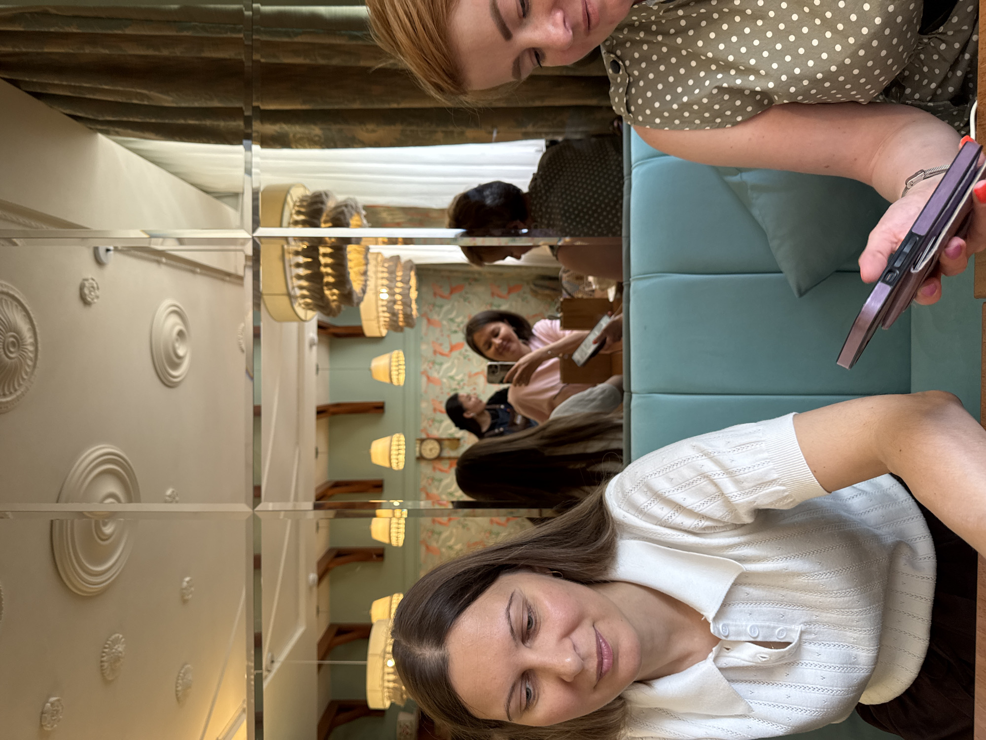 The image depicts a group of people in what appears to be a brightly lit interior space, possibly a cafe or restaurant, with mirrored walls creating a sense of depth and reflection. The orientation of the image is vertical, and the content is also reflected in the mirrors.
In the foreground, a woman wearing a white collared shirt is looking downwards and slightly to the right, with a gentle smile. Her long, brown hair is visible. To her left, a reflection shows another woman in a similar white shirt, also looking down.
Further into the reflected scene, another woman with dark hair is visible, holding a smartphone. She is wearing a pink top and appears to be interacting with the device. Behind her, a woman with blonde hair is reflected, wearing a green polka-dot top.
The mirrored walls reveal decorative elements of the room, including patterned wallpaper with birds, ornate ceiling moldings, and several hanging lamps with woven shades. The overall atmosphere seems relaxed and social, suggesting people are gathered for conversation or to enjoy the setting. The time of day is not explicitly indicated, but the lighting suggests it could be daytime or early evening. There is no visible text in the image. The location context provided is Izhevsk, Russia.