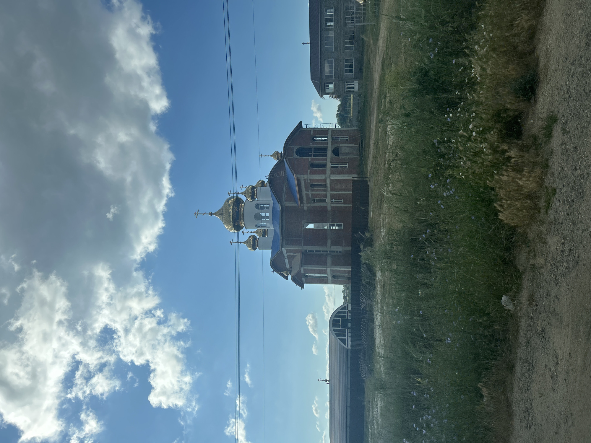 In the city of Oktiabrskiy, Russia, an unfinished church with golden domes stands prominently against a bright blue sky dotted with white clouds. The church is constructed of red brick, with its blue roof partially visible. Adjacent to the church, another building, seemingly residential, is visible. The foreground is dominated by a field of tall green grass and wildflowers, with a dirt road running along the bottom edge of the frame. Power lines are strung across the sky, and a fence is visible in the middle ground. The overall scene depicts a partially completed religious structure in a rural or suburban setting.