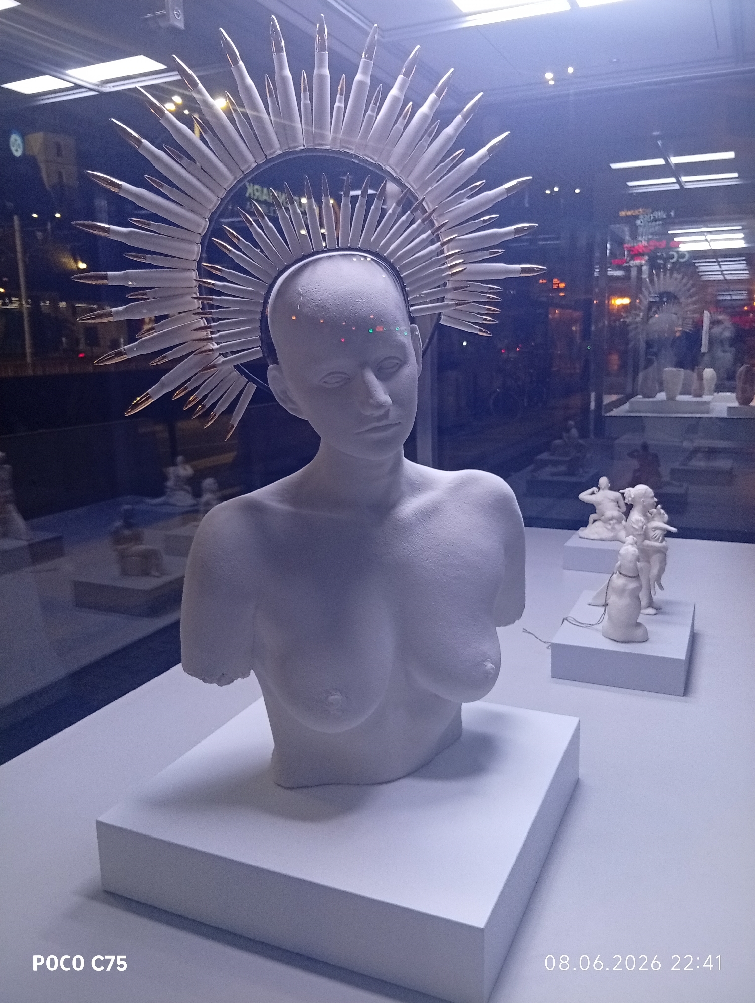 The image displays a white plaster bust of a headless female torso, adorned with a halo-like headdress made of white bullets with gold tips. The bust is positioned on a white pedestal, which sits on a white display surface. In the background, through a glass window, a city scene at night is visible, with blurred lights of buildings and vehicles, suggesting an urban environment. The overall presentation appears to be an art exhibition or display, possibly in a shop window, given the reflections and the arrangement of other small sculptures in the background. The timestamp "08.06.2026 22:41" and "POCO C75" are overlaid on the bottom right and left corners respectively. The reflection in the glass suggests the display is located in Wrocław, Poland.
