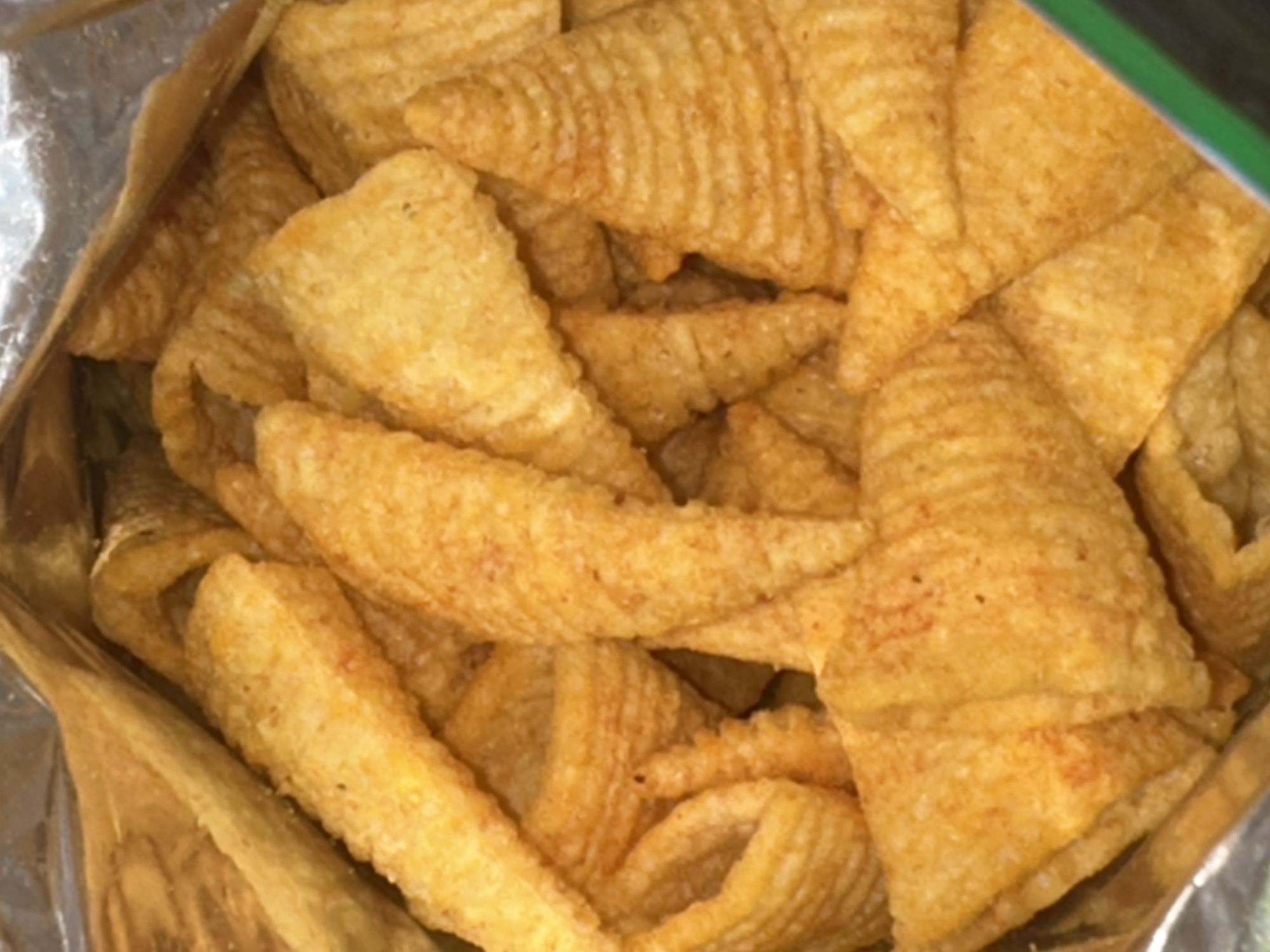 The image displays a close-up, overhead view of a bag of cone-shaped, ridged, corn-based snacks. The snacks are golden brown and appear to be seasoned. They are packed tightly together, filling the frame. The background is the silver interior of a chip bag, with a hint of green at the bottom right. The lighting is consistent, suggesting an indoor environment. There are no people or any identifiable city landmarks visible.