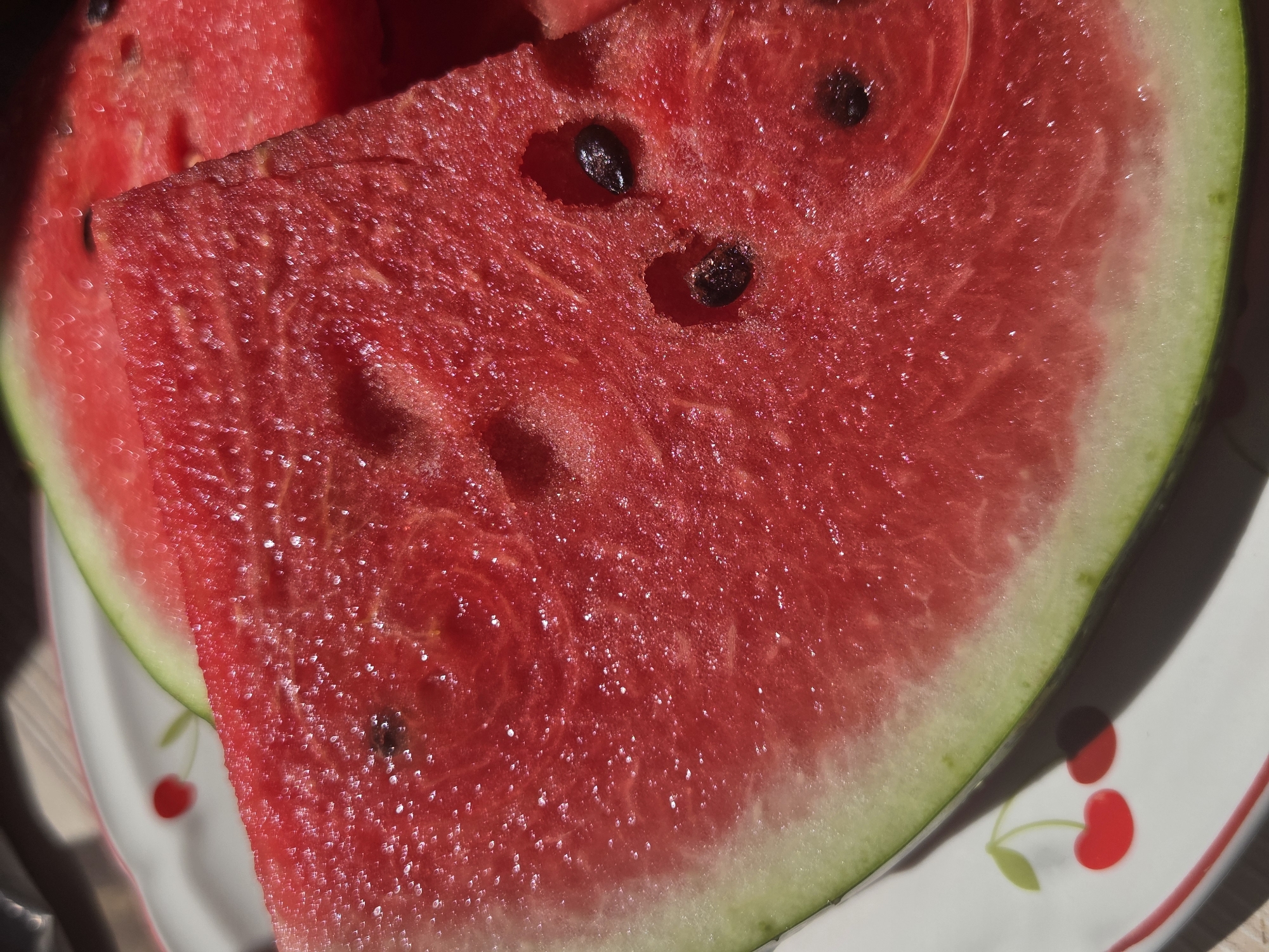 A close-up, overhead shot captures several slices of ripe watermelon on a white plate adorned with a cherry pattern. The watermelon flesh is a vibrant red, glistening with moisture and dotted with black seeds. The rind is a bright green. The scene is lit by natural light, casting shadows that highlight the texture of the fruit and the plate. The image appears to have been taken outdoors, possibly in a garden or patio area of a dacha in the city of Восход Снт (фролово), Russia.