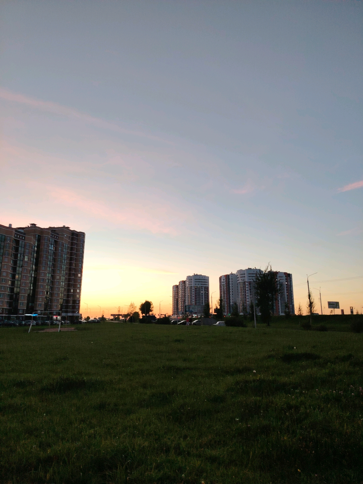 The image captures a view of residential buildings in a city, likely during sunset or dusk, given the gradient of colors in the sky. In the foreground, a vast expanse of green grass, possibly a park or open field, stretches across the frame. The grass appears to be natural and somewhat unkempt, with a few small white flowers scattered throughout.
In the middle ground, several multi-story apartment buildings dominate the scene. The building on the far left is tall and appears to be made of brick, with many windows. To the right of this building, a cluster of three modern-looking apartment buildings can be seen. These buildings are lighter in color and have a more contemporary architectural style, with visible balconies and windows. Some trees are interspersed between these buildings, and streetlights are visible. Cars are parked in front of some of the buildings and along a road that runs between the grassy field and the apartments.
The sky transitions from a pale blue at the top to warmer hues of orange and pink near the horizon, indicating either sunrise or sunset. The light is diffused, casting a soft glow over the scene. The overall impression is one of a suburban or developing urban area, with residential spaces adjacent to open green areas.
While the precise location within Vitsebsk, Belarus cannot be determined from the image alone, the architectural style of the buildings and the surrounding environment are consistent with urban landscapes found in Eastern European cities. The image depicts a peaceful evening scene, devoid of any prominent human activity or interaction.