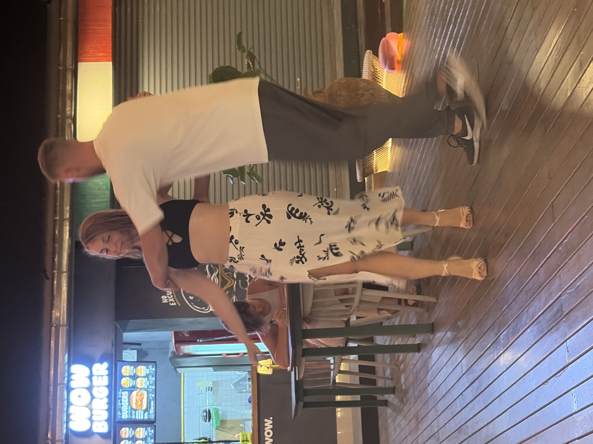 A man and a woman are dancing outdoors at night in what appears to be a restaurant or bar area. The man, wearing a white t-shirt and dark pants, is leaning over and holding the woman. The woman, in a black top and a white skirt with a black floral pattern, is looking down and smiling. She is wearing strappy sandals. The scene is lit by artificial lights, creating a warm ambiance. The ground is a wooden deck. In the background, there is a sign that reads "WOW BURGER" in bright blue neon letters. Behind the sign, a menu board with pictures of burgers is visible. There are also chairs and tables, suggesting a dining setting. The overall mood appears to be joyful and playful. The location is Ko Samui District, Thailand.