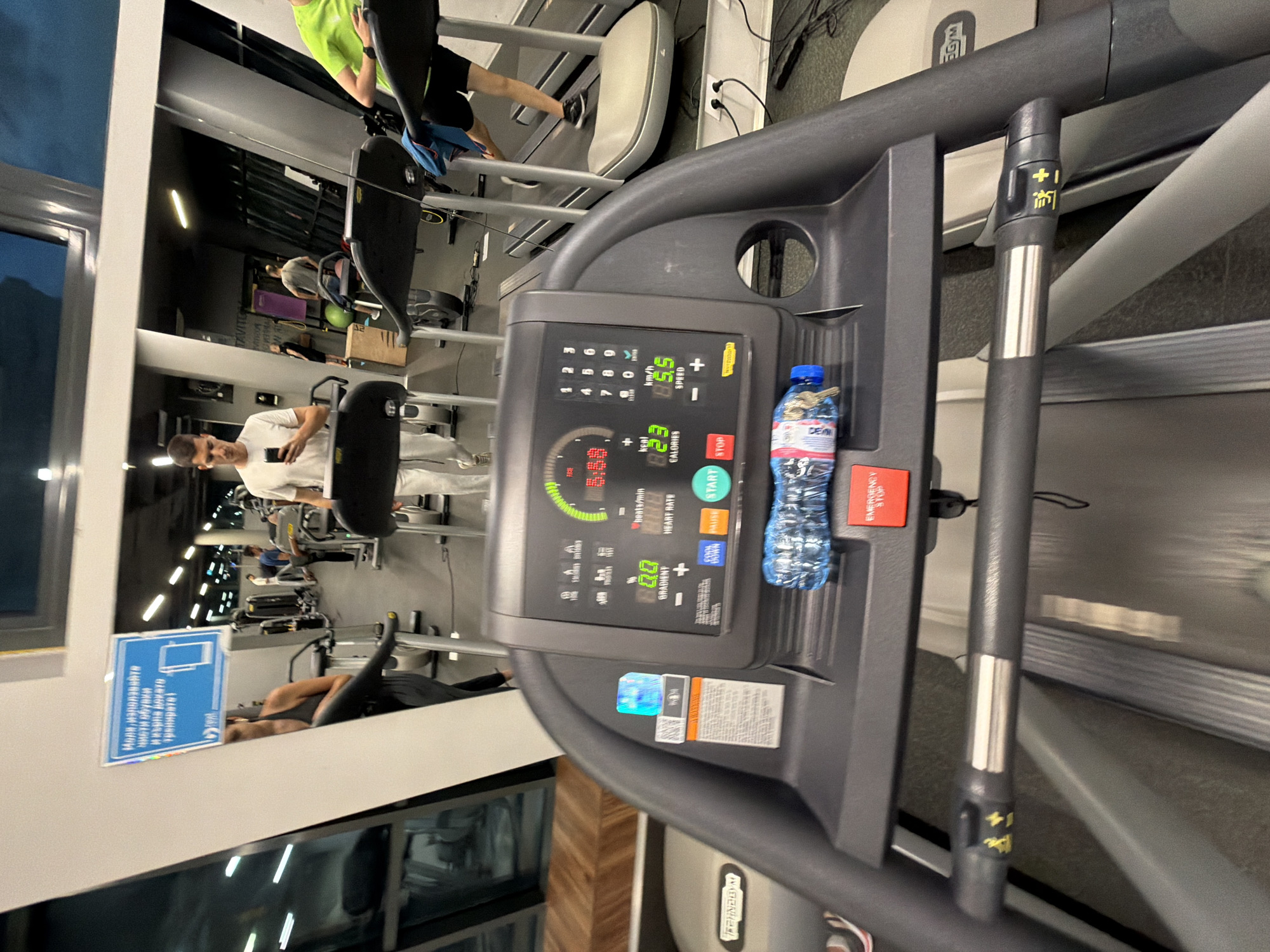 Here's a detailed description of the image:
**Setting:** The image appears to be taken inside a gym. The presence of treadmills, mirrored walls reflecting gym-goers, and gym equipment confirms this.
**Main Subject:** The primary focus is on the console of a treadmill. It displays various metrics such as speed (8.5 km/h), calories (82.3), heart rate, and gradient. There's a water bottle placed on the console.
**People:** Reflected in the mirrored wall, we can see multiple individuals. Some are on treadmills, others are using different gym equipment. One person is wearing a bright green shirt.
**Objects:**
* Treadmill console with buttons (Start, Stop, Pause, incline) and a display screen.
* Water bottle.
* Mirrors.
* Gym equipment (partially visible in the background).
**Environment:** The gym has artificial lighting. The mirrored walls create a sense of spaciousness and allow a view of other gym users. There is a blue sign, in Bulgarian, visible on a wall.
**Based on the sign in Bulgarian, the location is likely Sofia, Bulgaria.**