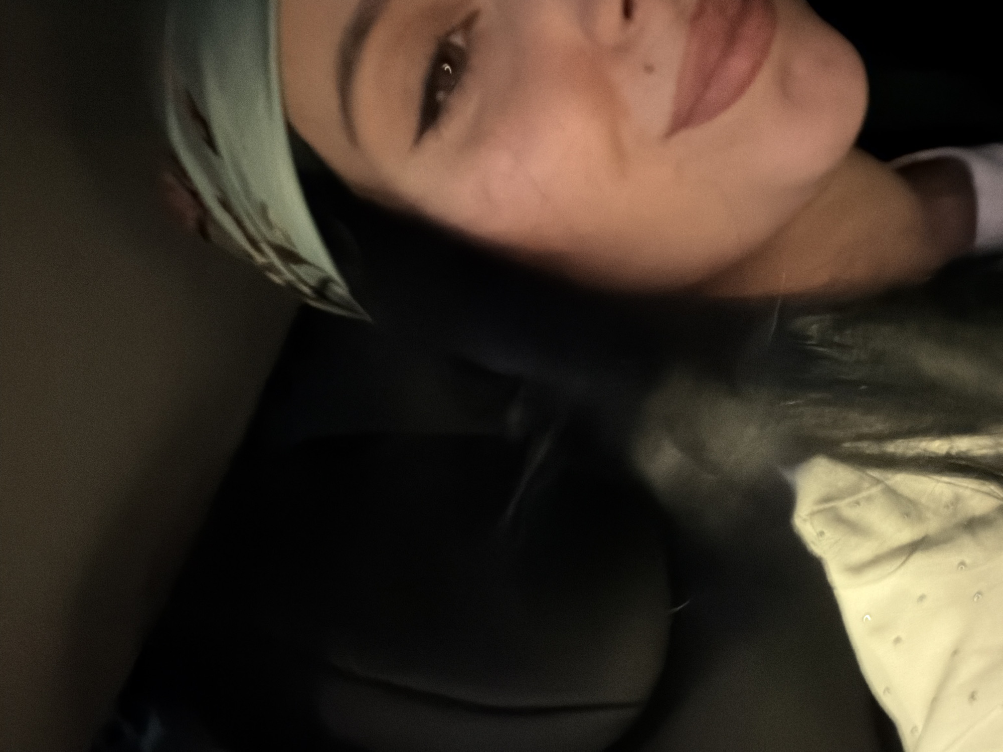 The image shows a close-up, angled selfie of a young woman, likely taken inside a vehicle. She has dark hair, is wearing a light blue headscarf with a patterned design, and has noticeable eyeliner. Her left eye and part of her mouth are visible, with a slight smile. She is wearing a light-colored shirt with small, light-colored dots. The background is dark and out of focus, appearing to be the interior of a car, with a dark seat visible on the left side. The overall lighting suggests it might be nighttime or a dimly lit interior. There is no indication of the location being in Tbilisi, Georgia.