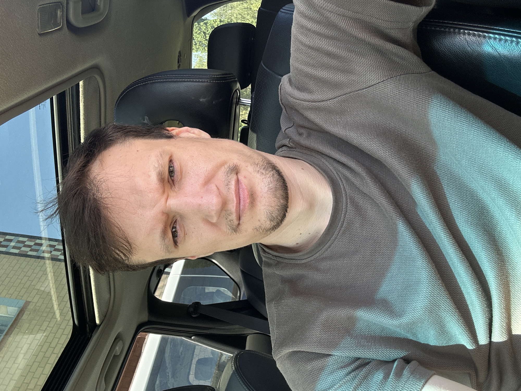 The image shows a man sitting in the driver's seat of a car with a sunroof. He has short brown hair, a light complexion, and is wearing a dark grey t-shirt. He is looking directly at the camera with a slightly furrowed brow and a neutral expression. The interior of the car is visible, including black leather seats and a portion of the dashboard. The sunroof is open, revealing a clear blue sky and the reflection of a brick building. The image appears to be taken during the daytime, with sunlight casting shadows.