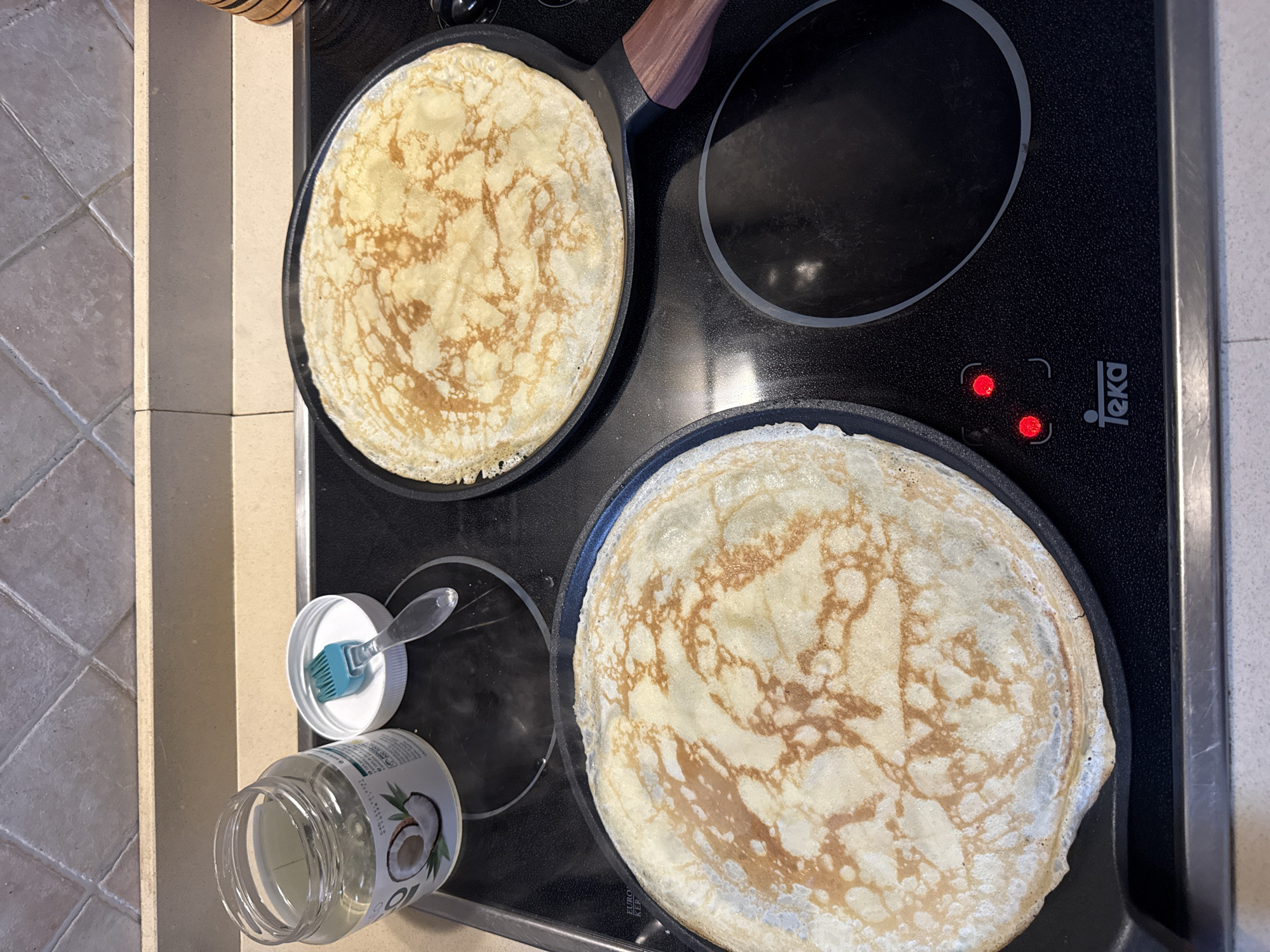 The image displays an overhead view of a kitchen countertop featuring two frying pans with cooked crepes on an induction cooktop. The cooktop, branded "Teka," is black and has two illuminated red indicator lights to the lower left. Two round, light golden-brown crepes, each exhibiting distinct browning patterns, are present in separate black frying pans. To the left of the cooktop, a partially full, clear glass jar of solidified coconut oil with a white label featuring a coconut image is visible. Its white lid, with a turquoise silicone basting brush resting on it, is placed beside the jar. The immediate countertop surface is a light, speckled material. Behind the countertop, a wall is covered in light brown square tiles arranged in a grid pattern. No individuals are present in the scene.