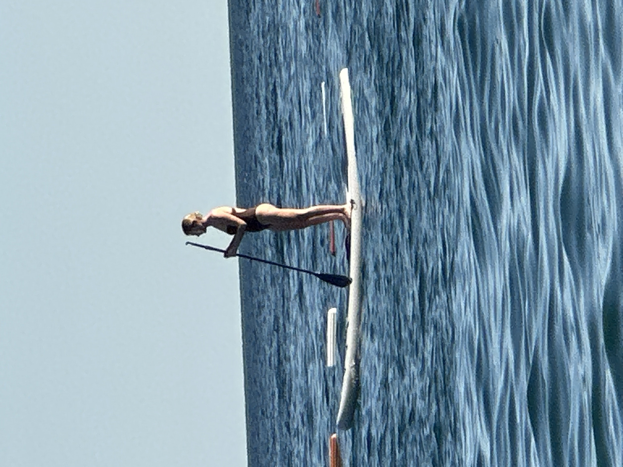 A woman stands on a paddleboard in the Adriatic Sea, off the coast of Boreti, Montenegro. The sky is a clear, pale blue. The sea is calm, with gentle ripples. The woman is wearing a dark swimsuit and holding a paddle. She is looking out at the water. In the background, there is a thin strip of land that marks the coastline. The image is shot from a slightly low angle, with the water taking up most of the frame.