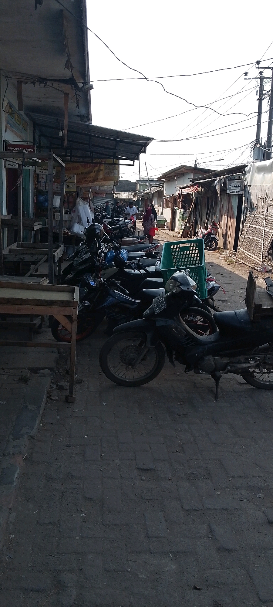 The image depicts a street scene in Sriamur, Indonesia, likely a market area. The foreground is dominated by several parked motorcycles, some with visible license plates. To the left, a wooden market stall structure is visible, with empty shelves and a concrete base. Further back, more motorcycles are parked, and people are walking. The background shows a row of small buildings and stalls with corrugated metal roofs and wooden walls. Electrical wires crisscross overhead, supported by utility poles. A green plastic basket is placed on top of a motorcycle. The ground is paved with interlocking concrete blocks. The lighting suggests it is daytime, possibly late afternoon, with the sun casting shadows.