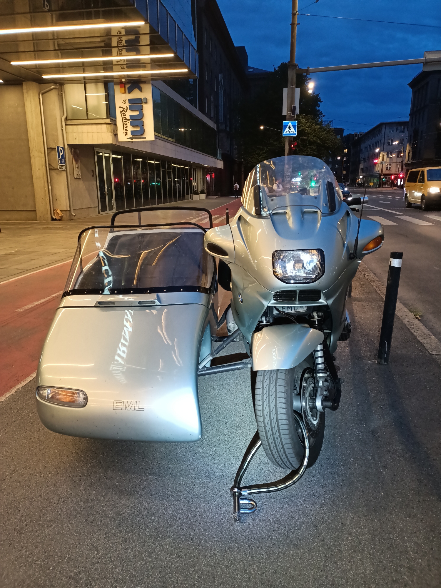 A silver motorcycle with a sidecar is parked on a city street at dusk. The motorcycle has a large windshield, a headlight, and a front wheel. The sidecar is attached to the right side of the motorcycle and has a windshield and a single headlight. The motorcycle and sidecar are locked together with a heavy-duty U-lock.
In the background, there is a modern building with illuminated signs, one of which reads "Radisson Inn". The building appears to be a hotel. The street is paved and has a red-colored bike lane running along the sidewalk. Traffic lights are visible in the distance, indicating an intersection. A yellow van is driving on the street. The sky is dark blue, suggesting it is twilight.
The scene appears to be in an urban environment, possibly near a hotel or commercial area. The presence of a hotel sign and the overall architecture suggest an urban setting in Tallinn, Estonia. The image captures a typical city street scene with a parked motorcycle and sidecar, locked for security. No people are clearly visible in a way that would indicate specific actions or interactions, but silhouettes of people may be present within the hotel or walking on the sidewalk. The overall impression is one of a quiet evening in the city, with the motorcycle and sidecar being the main focus.