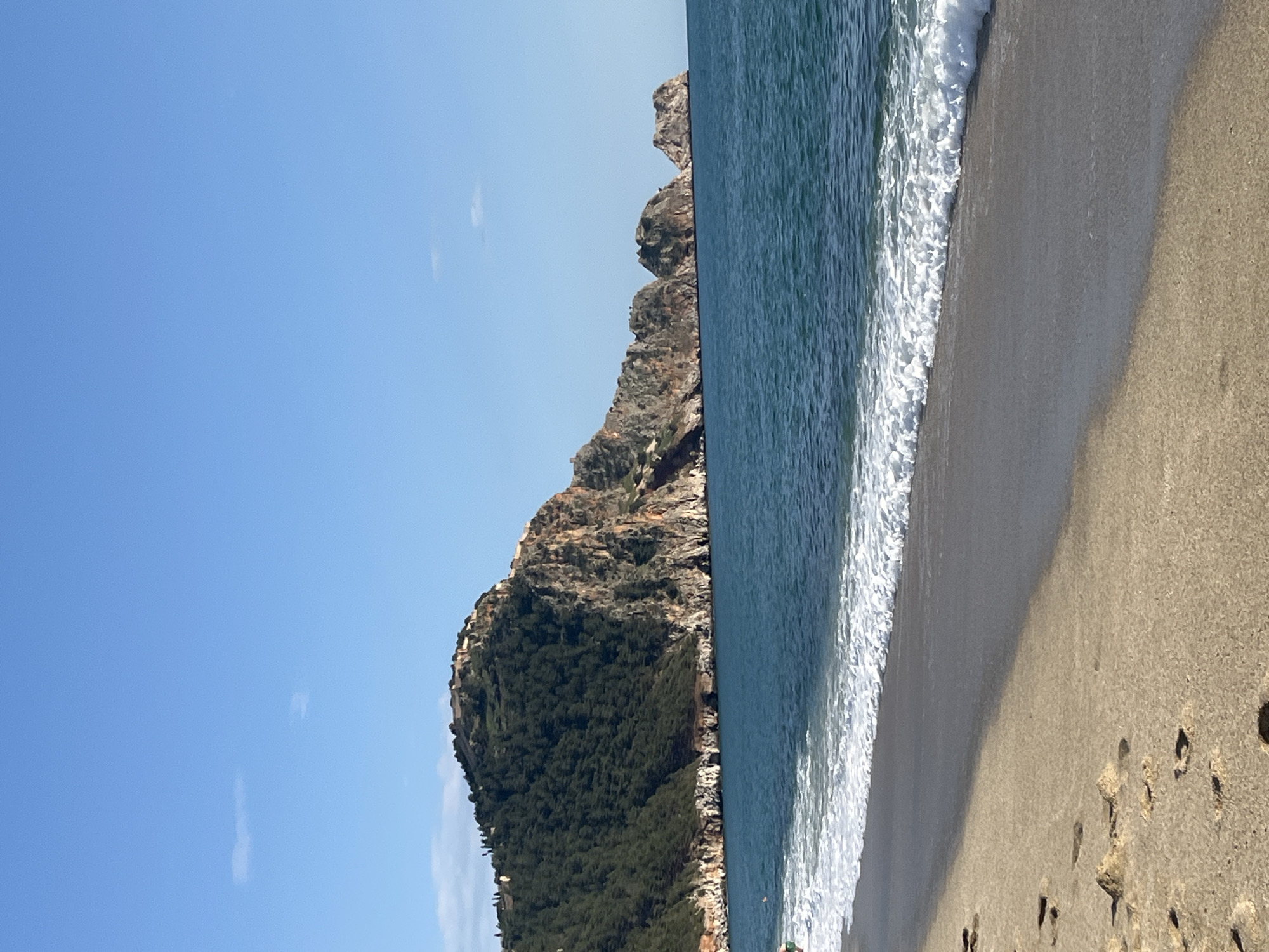 The image captures a beach scene in Alanya, Turkey. The clear, light blue sky is visible in the upper left quadrant. A rocky cliff covered in dense green vegetation runs along the center of the image, meeting the blue sea. White foamy waves break along the shoreline. A tan-colored sandy beach occupies the right side of the image, showing footprints. The scene appears calm and serene.