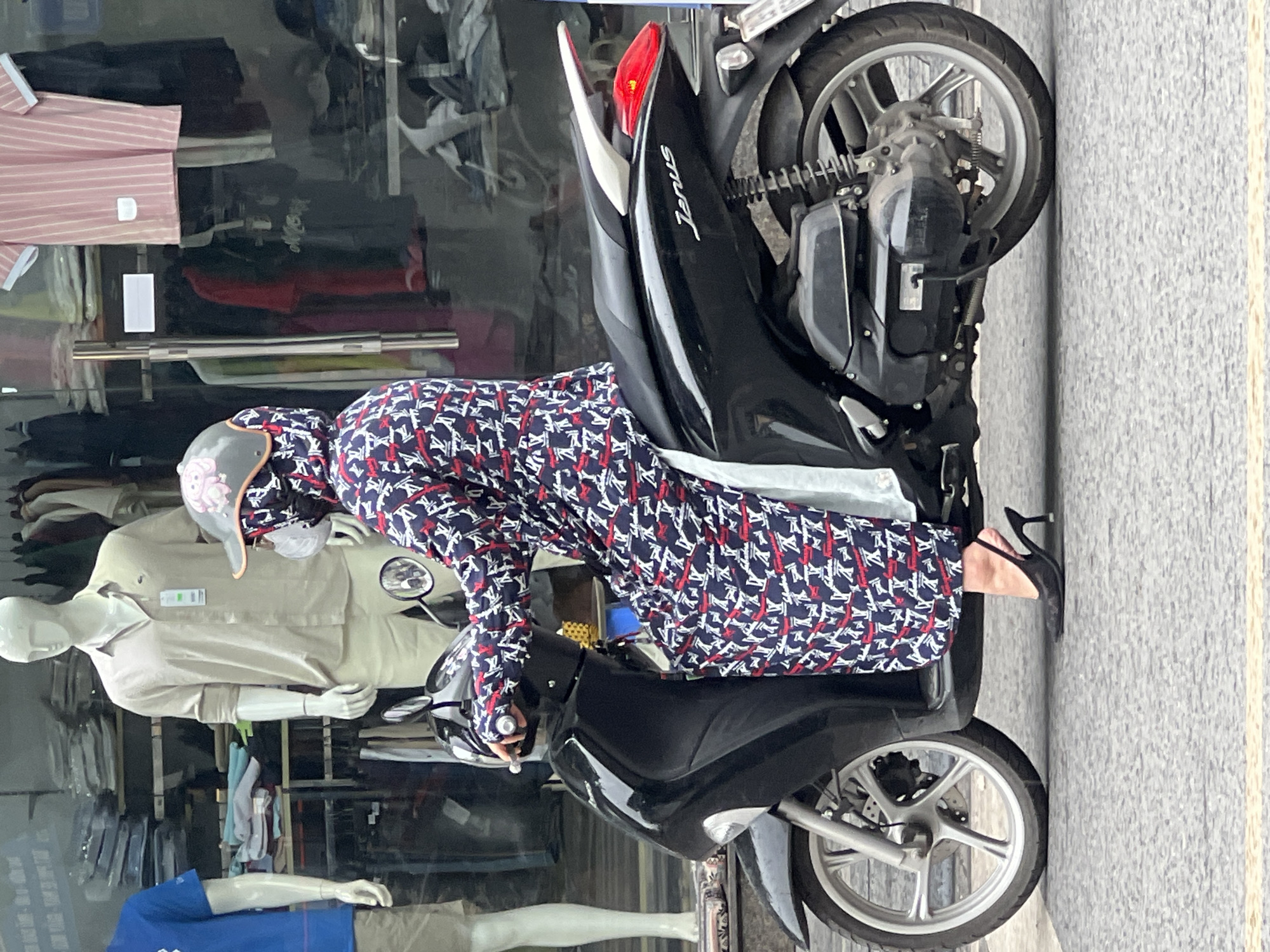 A woman wearing a face mask and a patterned long-sleeved jacket is riding a black scooter in a city. She is wearing high heels and a helmet adorned with a pink cartoon character. In the background, a store display features mannequins in casual clothing, and racks of garments are visible. The scene appears to be on a street in Hanoi, Vietnam, with the pavement and storefront contributing to the urban setting.