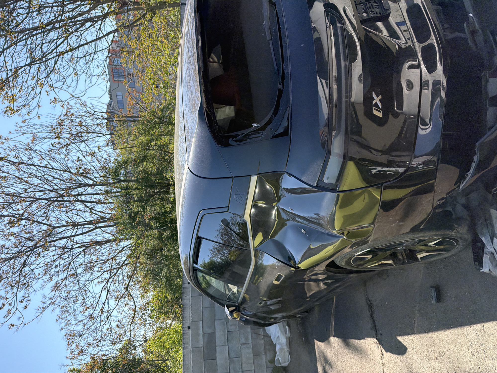A black BMW iX SUV with significant damage to its front passenger side is parked on a paved surface next to a low concrete wall. The damage includes a crumpled fender and compromised front panel. The setting appears to be outdoors, possibly a residential area, indicated by trees and a building in the background. The sky is clear, suggesting daylight. The SUV bears the "iX" model designation. The image provides no further detail to establish the location in the city of Burgas, Bulgaria.