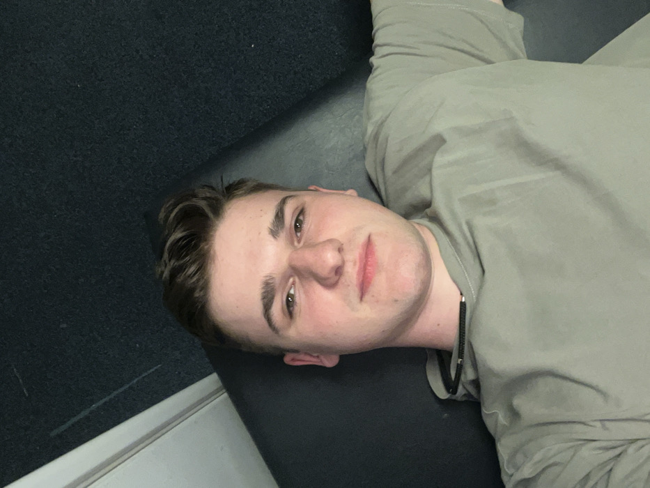 An indoor photograph, purportedly taken in Gafanha da Nazaré, Portugal, captures a single young male in a head-and-shoulders portrait. The subject, an individual estimated to be between 18 and 25 years old, possesses short brown hair, a light complexion, and brown eyes. He is wearing a light olive-green short-sleeved top, possibly a t-shirt or lightweight hoodie. A thin, dark-colored cord or braided necklace with minor metallic accents is visible around his neck, resting just above the collar. The male is positioned in a supine or reclining posture on a dark, upholstered surface, with his head supported. His gaze is directed forward, toward the camera. The background is uniformly dark, consistent with a dark grey or black carpeted floor or wall, exhibiting a faint light vertical line on the far left side of the frame, possibly indicating a wall edge or doorframe.