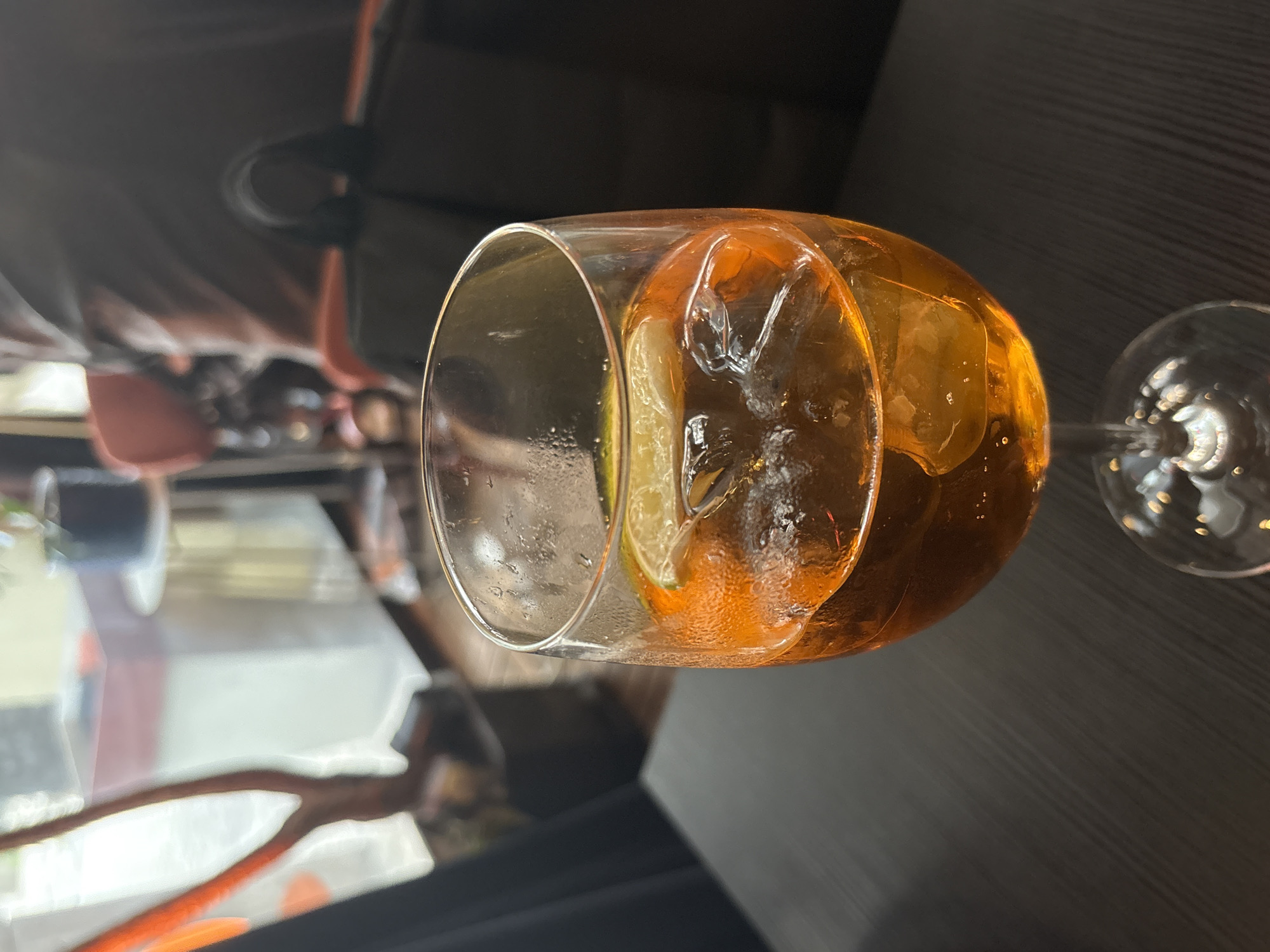 The image depicts a glass of amber-colored liquid, likely an Aperol Spritz, containing ice and a slice of lime. The glass is positioned on a dark, horizontally-textured surface, possibly a table. The glass has condensation on its exterior. The background is out of focus, but hints at an indoor restaurant setting, evidenced by the presence of table settings, chair backs, and partial views of other patrons. Given the prompt, it is reasonable to assume the photograph was taken in Belgrade, Serbia. No people are interacting directly with the beverage in the frame.