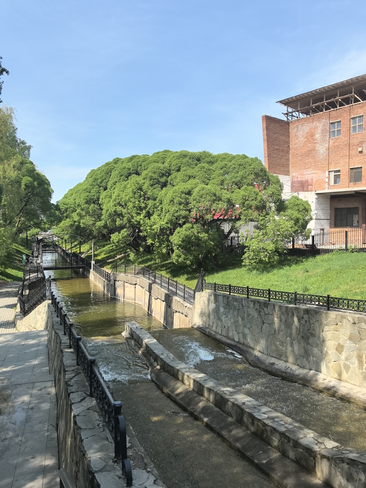 The image depicts a canal in the city of Perm, Russia. The canal is lined with stone walls and ornate black metal railings. Water flows through the canal in two channels. A walkway runs alongside the canal on the left side of the image. Lush green trees line the banks of the canal, partially obscuring buildings and structures in the background. The sky is clear and blue. A red brick building with an unfinished roof extension is visible on the right side of the canal.