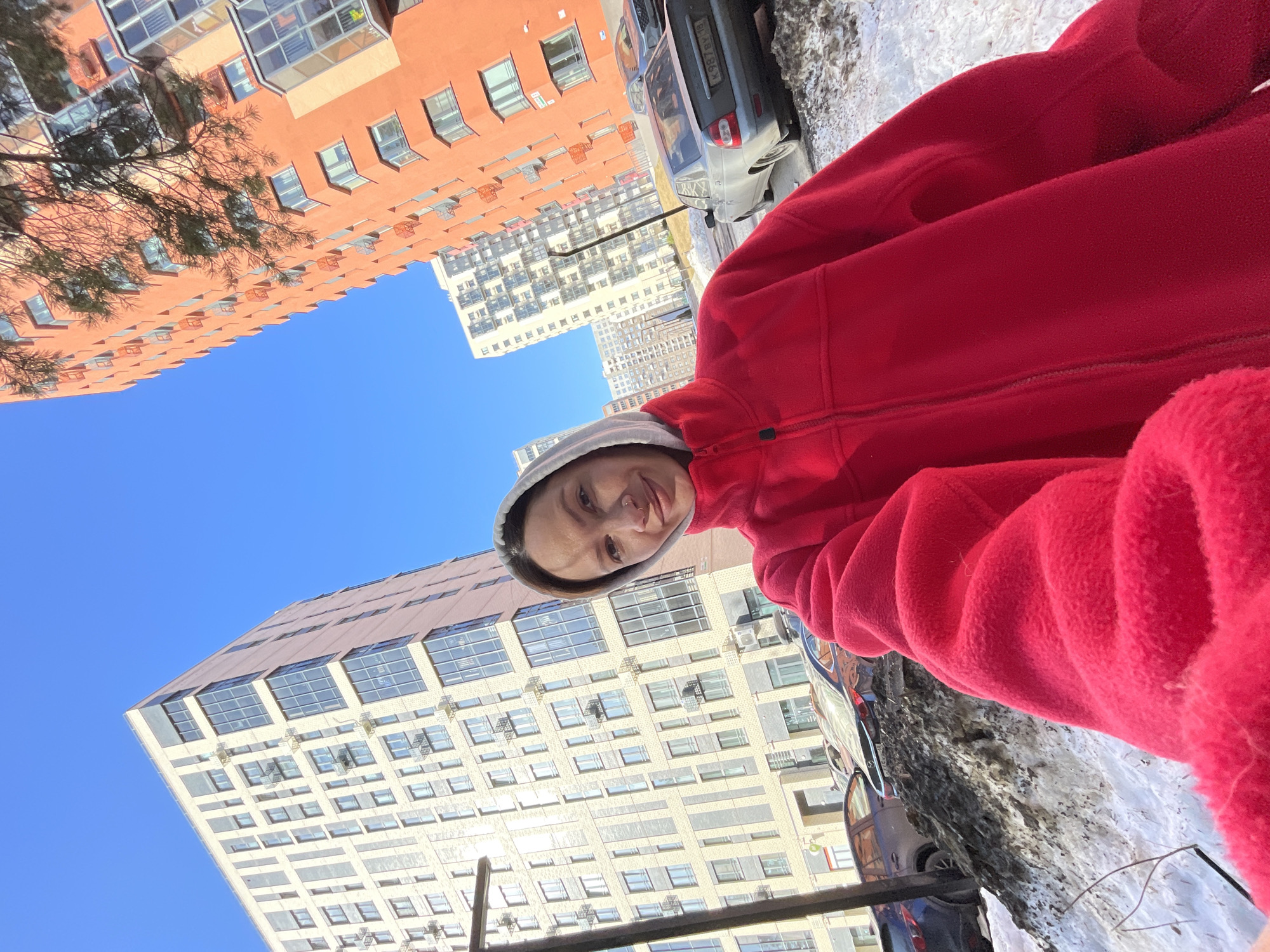 A selfie of a woman in a red fleece jacket and grey hoodie. She's standing outside in what appears to be winter conditions with some snow visible on the ground. In the background are several tall apartment buildings under a clear, blue sky. Some parked cars are visible. The photograph appears to have been taken in Izhevsk, Russia.