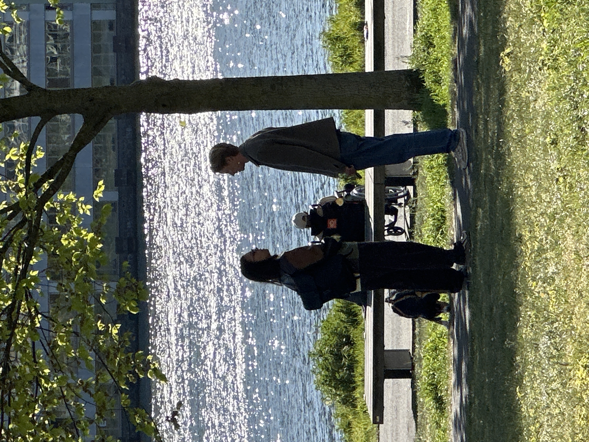 This outdoor photograph, taken in Amsterdam, Netherlands, captures a sunny moment by a sparkling body of water, likely a canal or river. The image is oriented vertically but depicts a horizontal scene.
In the midground, two individuals stand on a paved pathway bordered by short green grass. To their left (when viewed correctly), a large body of water reflects intense sunlight, creating numerous bright sparkles. On the far left, a substantial tree trunk rises, with leafy branches extending into the upper left quadrant, partially obscuring a building across the water.
The individual closer to the water (on the left side of the corrected image) wears dark clothing, including a dark jacket and pants, and appears to be carrying a dark backpack. Adjacent to them is what looks like professional camera equipment on a tripod, suggesting they might be involved in photography or filming. This person is looking slightly to their right, away from the camera.
Further to the right on the pathway stands another individual, wearing a lighter-colored jacket, blue jeans, and light-colored sneakers. They have light-colored hair and are also looking downwards and slightly to their right.
In the background, across the shimmering water, a modern building with visible window panes can be faintly seen through the tree branches. The bright reflections on the water and the shadows cast on the grass indicate a clear, sunny day. The scene conveys a sense of quiet observation or preparation in an urban waterfront setting. No visible text is present in the image.