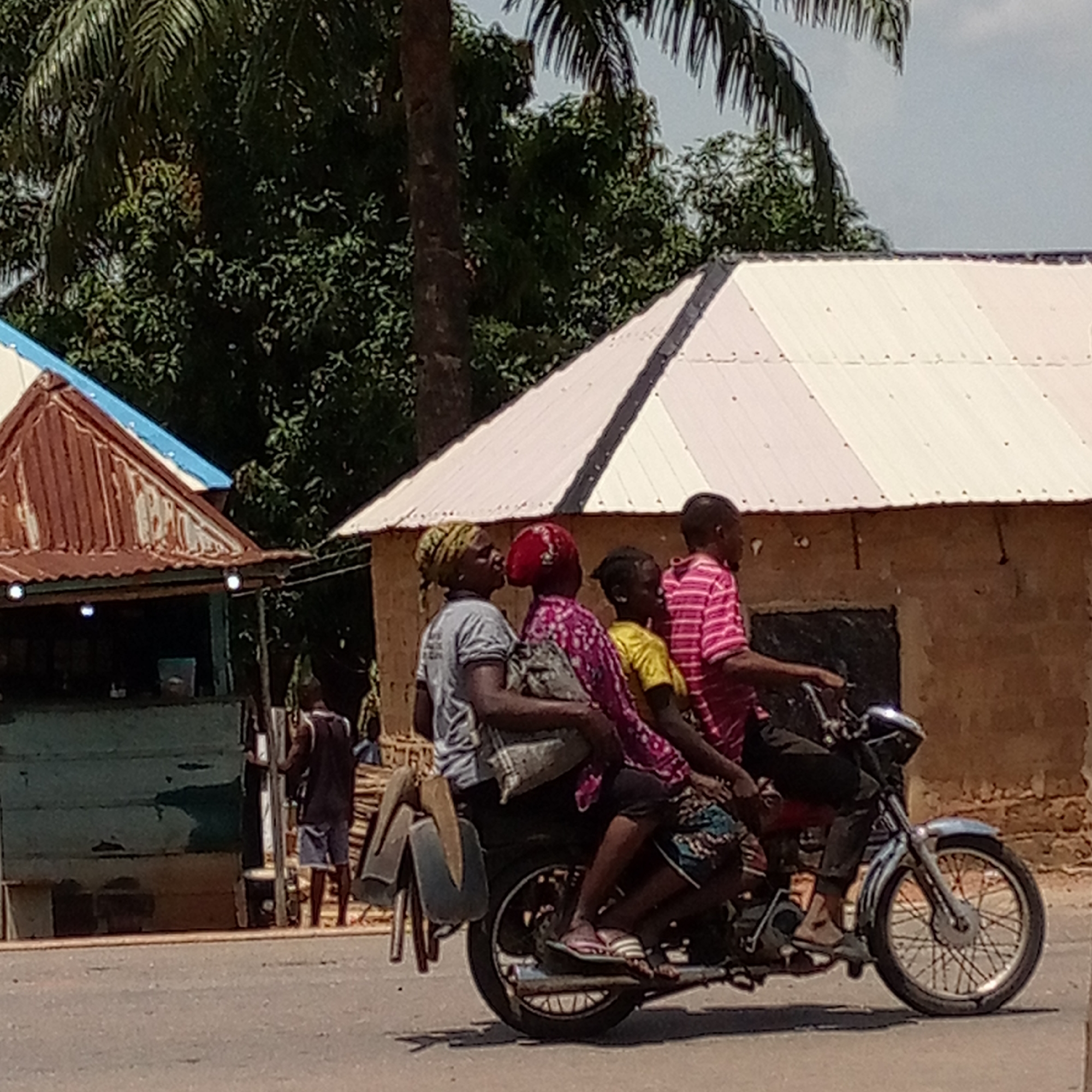 The image depicts a motorcycle with four people riding on it, traveling along a paved road in Mararaba, Nigeria. The rider, a male wearing a pink striped shirt and dark pants, is operating the motorcycle. Behind him are three passengers: a woman in a gray shirt and headwrap, a woman in a pink patterned shirt, and a child in a yellow shirt. A building with a white and dark striped roof is situated to the right of the motorcycle, and a smaller structure with a rusty metal roof is visible to the left. A man is standing near the smaller structure. A palm tree and other trees form the backdrop.