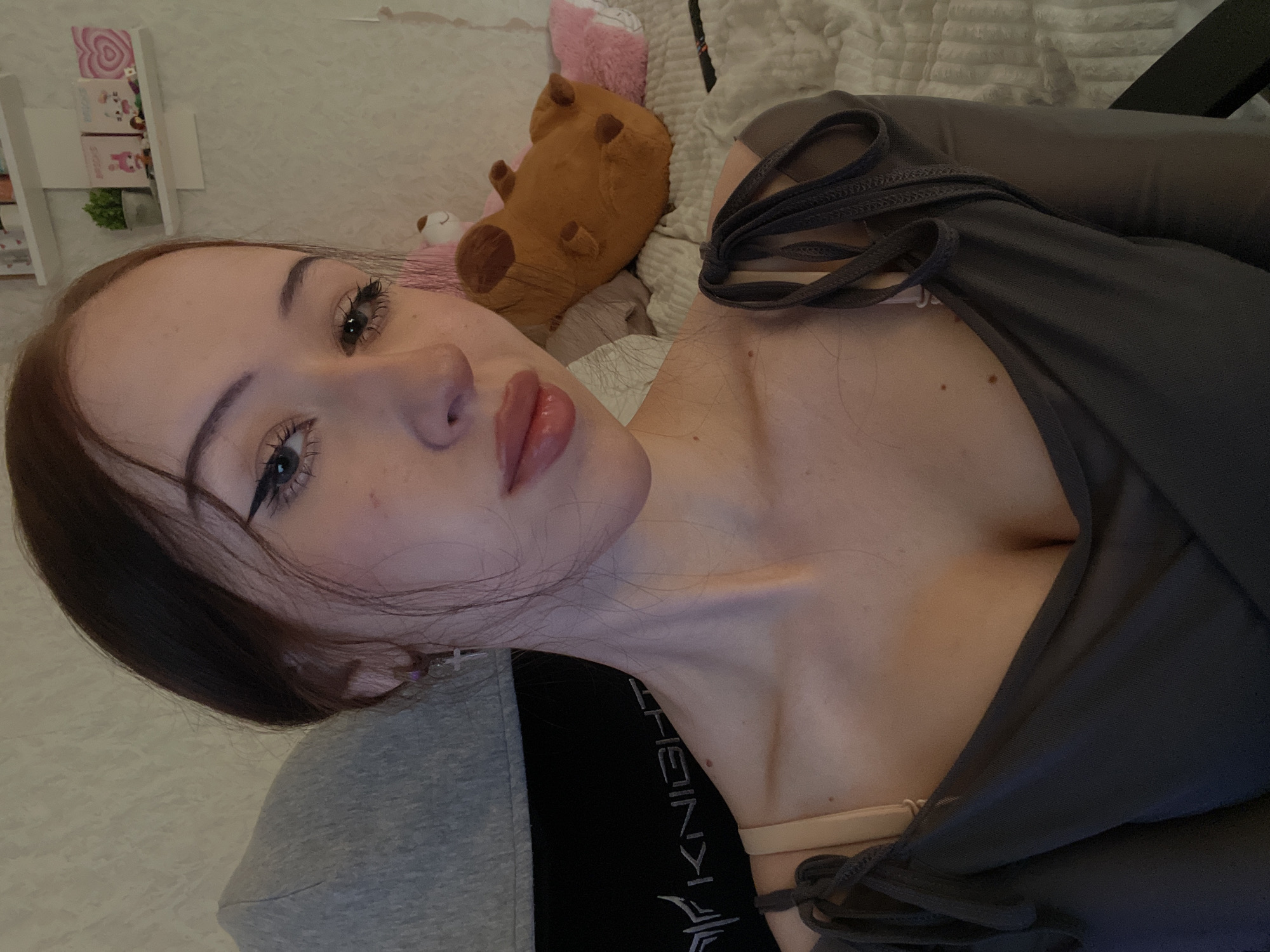 The image depicts a young woman posing for a selfie in an indoor setting, identified as being in the city of Votkinsk, Russia. The woman, with dark hair parted on the side and some strands framing her face, looks directly at the camera with a neutral expression and slightly parted, glossy lips. She has visible eyeliner with a winged style and dark mascara on her eyelashes. A small cross earring hangs from her left ear. She is wearing a dark, possibly grey or charcoal, off-the-shoulder or tie-strap top, and a beige bra strap is visible on her right shoulder. A small mole is present on her upper chest, below her right collarbone.
The background reveals a domestic environment. To the left of the woman, a grey and black gaming chair is partially visible, with the word "KNIGHT" printed in white on the black portion. To her right, a light-colored wall with a subtle textured pattern is present. On the wall, two white floating shelves hold various decorative items, including a small potted plant, framed pictures or art pieces (one featuring a pink heart), and small figurines. Behind the woman, to the right, a bed with a light-colored, textured duvet or blanket is visible. On the bed, a large brown plush toy, possibly a dog or bear, is positioned alongside a pink pillow. A dark-colored object, potentially a remote control, rests on the bed near the pillow.