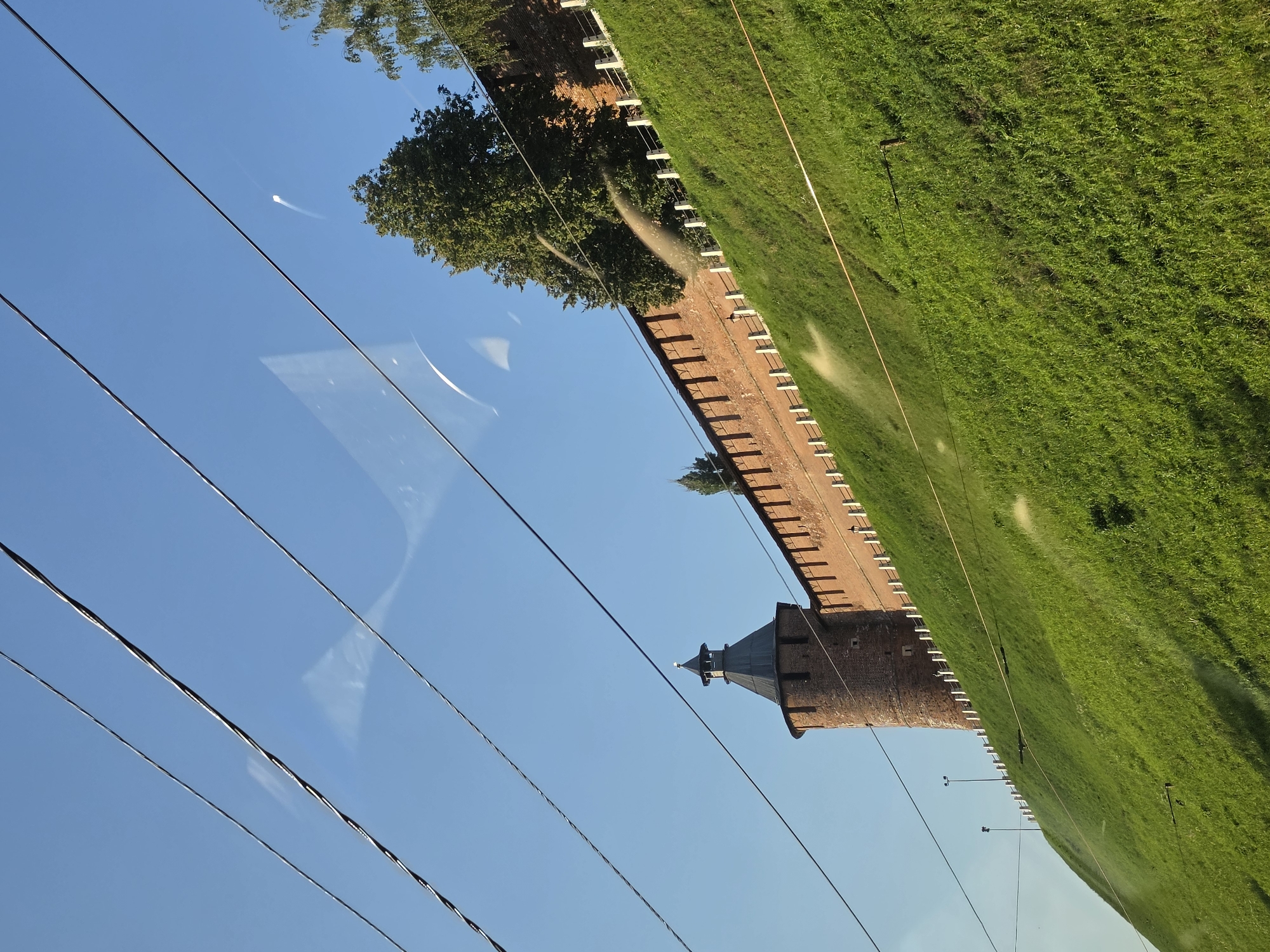 The image captures a view of the Nizhny Novgorod Kremlin's defensive wall and a round brick tower, seen from a lower vantage point and partially obscured by overhead wires. The Kremlin wall, constructed of red brick, stretches diagonally across the frame from the upper right to the mid-left. A round, dark-capped tower with a pointed roof stands prominently to the left of the wall. Both structures are situated atop a steep, lush green grassy slope. White posts with railing are visible along the edge of the slope just below the Kremlin wall. The sky is a clear, bright blue. There are visible water sprays on the grass, suggesting an irrigation system is in operation. The scene appears to be an outdoor, daytime view, likely captured from a moving vehicle, given the presence of the wires and the perspective.
