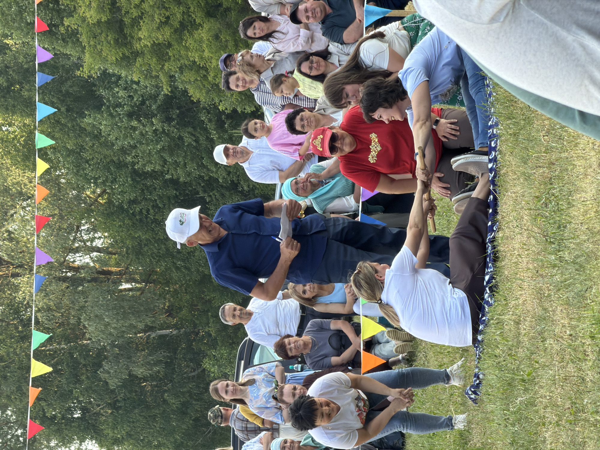 An outdoor gathering is taking place with a crowd of people of various ages watching a competition. Two women are seated on a patterned blanket on the grass, facing each other and gripping a wooden stick. Their hands are clasped around the stick, and they are engaged in what appears to be a test of strength or a game. The woman on the left is wearing a white t-shirt and blue jeans, and the woman on the right is wearing a light blue t-shirt and jeans.
In the background, a man in a blue shirt and white cap is holding a piece of paper and appears to be officiating or announcing. Other spectators are standing and sitting around, some are wearing casual attire, and some children are present. Colorful triangular flags are strung across the top of the frame, suggesting a festive occasion. The environment is grassy with trees visible in the background, indicating an outdoor park or recreational area. The lighting suggests it is daytime, likely late afternoon due to the warm tones.