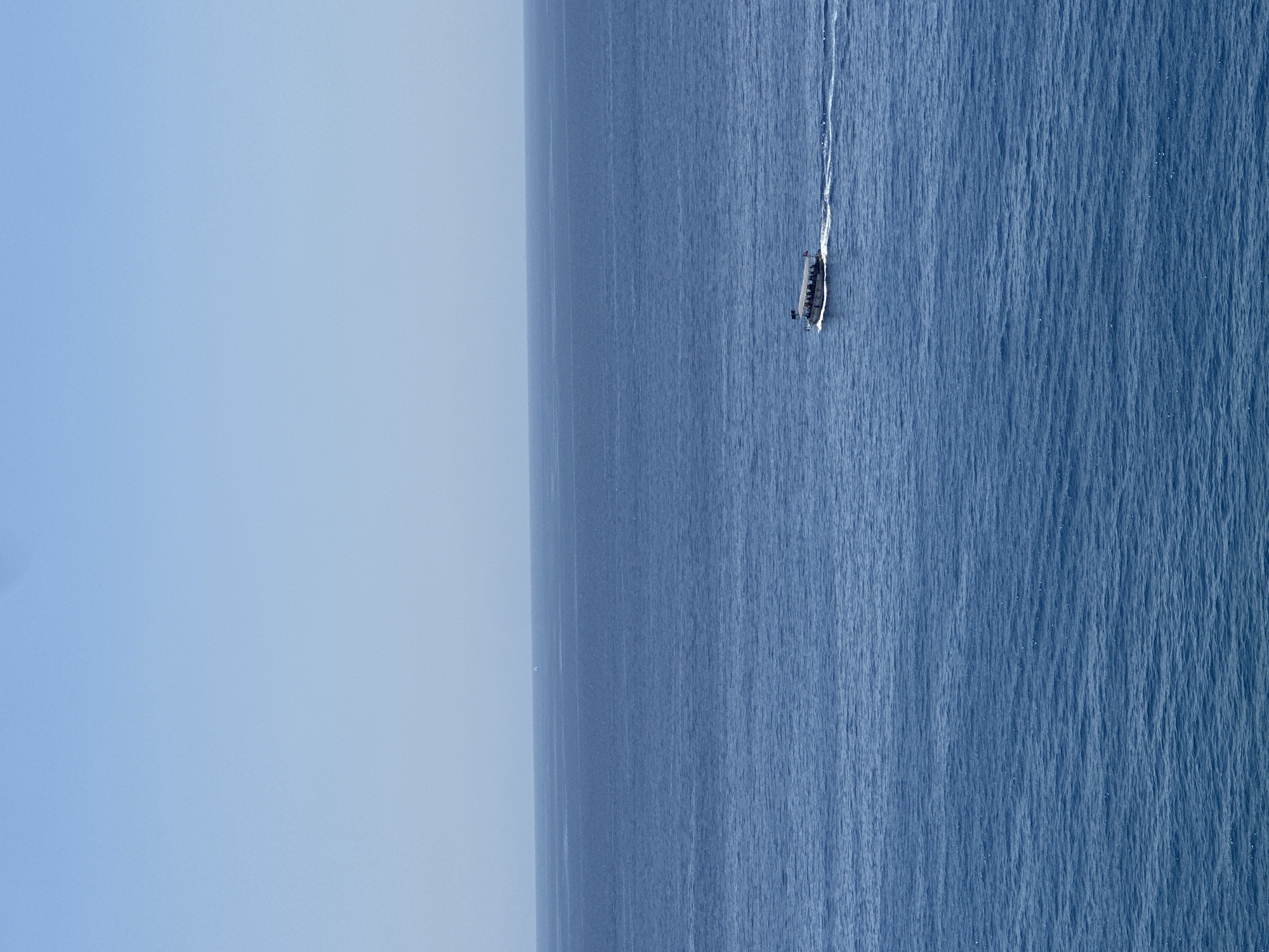 A motorboat carrying passengers travels across a vast expanse of blue ocean under a clear sky. The boat is positioned in the right-center of the frame, leaving a white wake behind it. The horizon line, separating the sea and sky, is visible in the upper portion of the image. The water exhibits gentle ripples.