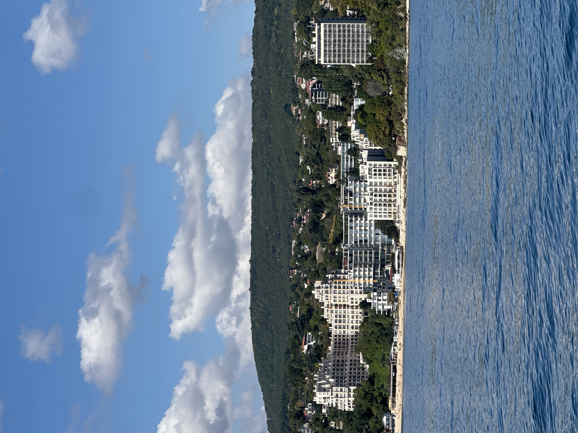 The image shows a coastal city with a body of water in the foreground and buildings and a hill in the background. The water is a deep blue with gentle ripples. Along the shoreline, there is a sandy beach with people sunbathing and some umbrellas set up. Behind the beach, a line of white buildings rises up, some appearing to be hotels or apartment complexes, with multiple stories and balconies. Further inland, a densely forested hill forms the backdrop, with some houses visible nestled among the trees. The sky is blue with scattered white, fluffy clouds. The overall scene suggests a sunny day at a seaside resort.