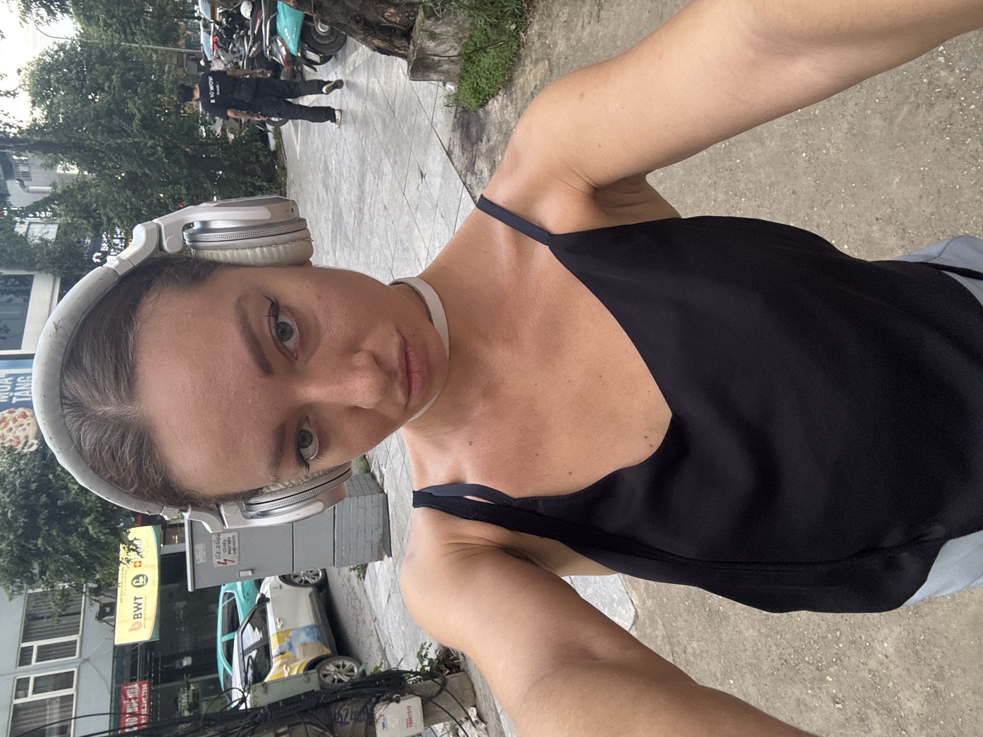A young woman with light brown hair, wearing large silver headphones and a black tank top, takes a selfie outdoors in what appears to be an urban setting in Hanoi, Vietnam. She is positioned in the foreground, with her face angled slightly towards the camera, and her left arm is extended to capture the shot. The background shows a street with a tiled sidewalk, a building with windows, a white car with a yellow advertisement on its side, a turquoise car, a metal utility box with signage, and parked motorcycles. A man in a black t-shirt and dark pants is visible walking away from the camera in the background. Trees and greenery are also present, suggesting an outdoor, possibly pedestrian-friendly area. The lighting indicates it is daytime, with overcast skies.