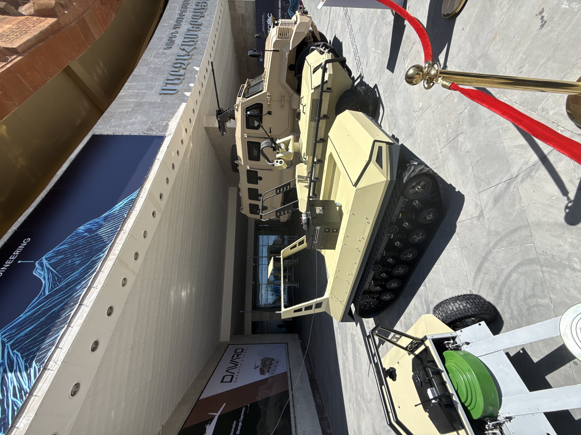 An outdoor scene displays several military or defense vehicles and equipment, likely at an exhibition or demonstration, marked off by red velvet ropes and gold stanchions on a paved surface. The image is captured in bright daylight, casting sharp shadows.
The primary subject is a large, tan-colored military-style vehicle, possibly an armored personnel carrier (APC) or a tactical vehicle, featuring multiple windows, rugged tires, and what appears to be a heavy machine gun mounted on a turret on its roof. Next to it, to the right, is a smaller, tan-colored tracked vehicle, possibly an unmanned ground vehicle (UGV) or a specialized tracked platform. Further to the right and partially visible, there is a trailer with pneumatic tires and a large, bright green reel or spool mounted on its frame.
In the background, a modern building with a light-colored facade rises. Prominently displayed on the building's upper section is text in an unfamiliar alphabet, which reads "ՄԻԱՍՆԱԿԱՆ ԼՈՒԾՈՒՄՆԵՐ" (Armenian for "Unified Solutions" or "Integrated Solutions"). Below this, a large blue banner features an abstract, glowing blue design and partially visible text "ENGINEERING." Beneath the blue banner, another white banner is visible, bearing the logo and text "DAVARO DEFENCE SYSTEMS," accompanied by the slogan "Innovation Strategic Supremacy." Adjacent to the Davaro banner, a smaller banner shows the letters "CMC." The overall impression is a professional display of defense technology.