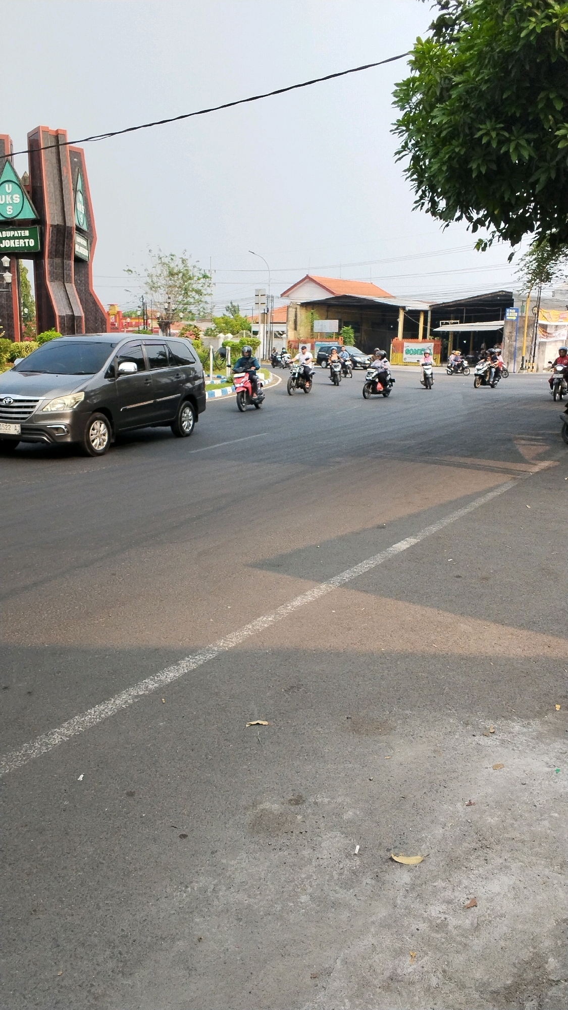 This image depicts a street scene in Banjaragung, Indonesia, near a landmark structure that appears to be a monument or a gateway, possibly marking the entrance to the district. The monument has a distinctive architectural design with multiple vertical elements, some of which are inscribed with text. In front of the monument, there is a dark gray SUV, a Toyota Innova, with its license plate visible.
The street is asphalted and divided by white lane markings. A procession of motorcycles is navigating the road. Several individuals are riding on motorcycles, some with passengers. The riders and passengers are wearing casual clothing, and some are wearing helmets. The traffic appears to be moving in the same direction.
In the background, there are buildings with orange roofs and awnings, suggesting a commercial or residential area. Trees with green foliage are visible, and the sky is overcast with a pale, diffused light, indicating it might be morning or late afternoon. The overall impression is of a typical day in a moderately busy urban or semi-urban environment in Indonesia.