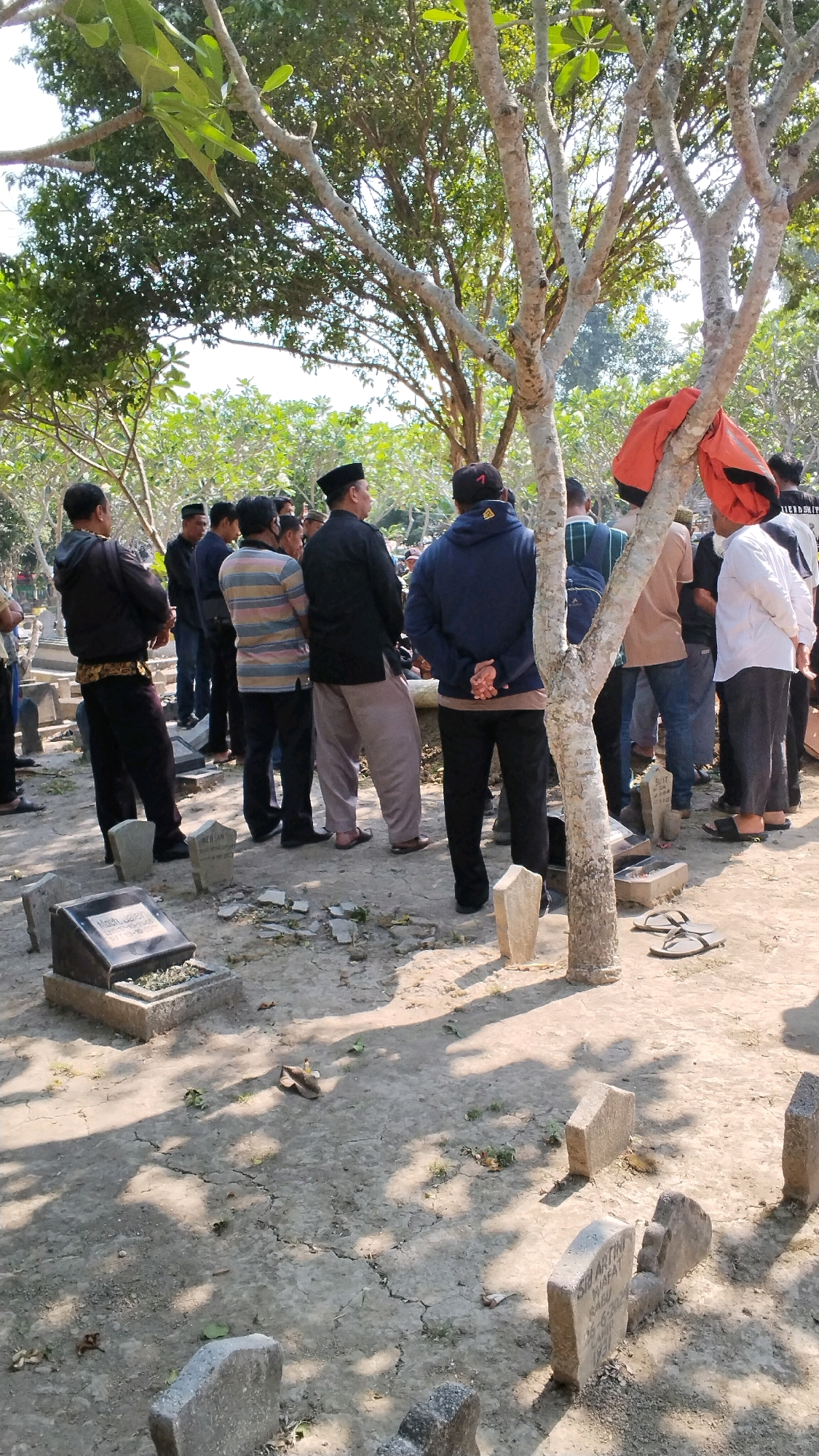 The image shows a group of people gathered in a cemetery in Sidoharjo, Indonesia. The people are of various ages and genders, dressed in traditional Indonesian attire. They are standing around a grave, some with their heads bowed in prayer or reflection. The cemetery is overgrown with grass and trees, and the graves are marked with simple headstones. The sun is shining brightly, casting long shadows across the ground. The overall mood of the scene is somber and respectful. The location in Sidoharjo, Indonesia, is identified by the style of the headstones, the clothing of the people, and the surrounding vegetation.