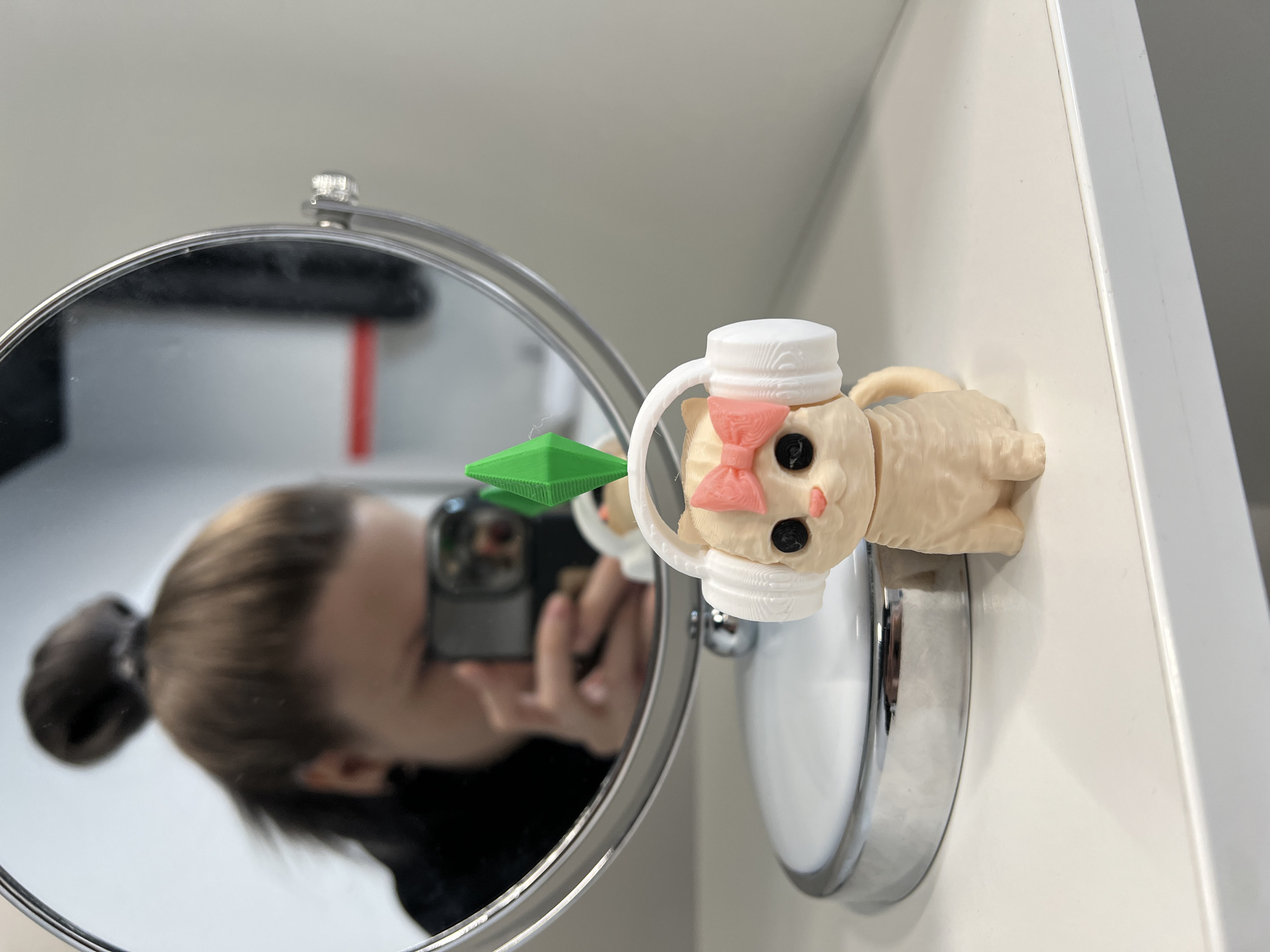 The image captures an indoor scene, likely a bathroom or vanity area, centered around a round, magnifying mirror with a chrome frame.
The primary subjects include a small, light-colored 3D-printed figurine resembling a cat or animal character. This figurine is attached to the white wall next to the mirror. It wears a white, cylindrical cap or hat with a handle-like projection, and a pink bow on its head. The figurine has black, round eyes and a small pink nose or mouth area. Above the figurine, and partially reflected in the mirror, is a vibrant green, pyramid-shaped object, possibly a "plumbob" accessory from The Sims game series.
Reflected prominently in the mirror is a person, likely female, with dark hair tied in a bun. The person is holding a smartphone, with its camera lens clearly visible, actively taking a picture of the scene. Their face is largely obscured by the phone.
The activity taking place is photography, with the person capturing an image of the figurine and its reflection in the mirror. The background consists of a plain white wall and the edge of a white cabinet or shelf, indicating a clean and modern environment.
Notable details include the texture of the 3D-printed figurine, which shows distinct layer lines. The lighting is bright and even, suggesting daytime, though the exact time and weather are not discernible. No visible text is present in the image. The location context provided is Khimki, Russia.