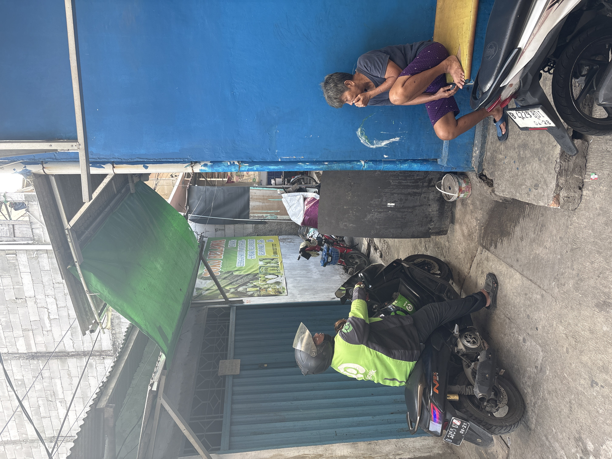 In North Jakarta, Indonesia, a street scene unfolds with two individuals. On the left, a person wearing a bright green and black uniform, complete with a helmet, sits on a black motorcycle. The motorcycle has a license plate that reads "B 3355 UTZ" and "08-23". The person appears to be stationary, possibly waiting. To the right, an elderly individual with short gray hair is crouched on a yellow bench against a vibrant blue wall. This person is wearing a dark t-shirt and purple shorts, with one hand touching their mouth. In the background, a store displays a sign with text in Indonesian, and various structures with corrugated metal roofs are visible, suggesting a densely built urban environment. The ground is paved with concrete, and a white bucket sits near the blue wall. Another motorcycle is partially visible in the foreground on the right.