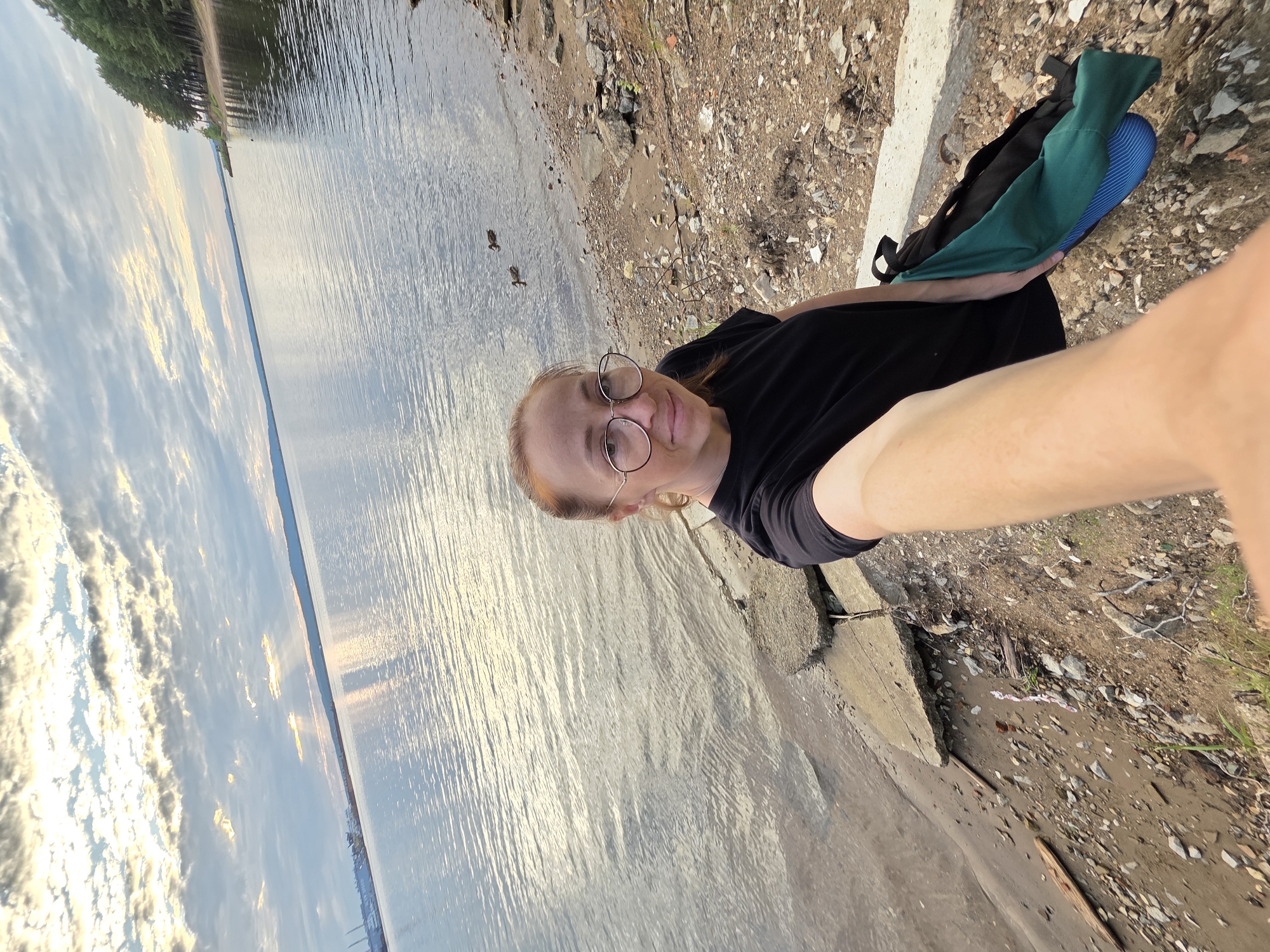 A person is taking a selfie on a shoreline. They are wearing a black t-shirt and round glasses, with their blonde hair pulled back. A backpack with a green cloth and blue item is on their right. The person is smiling at the camera.
The scene is a body of water, likely a lake or river, with a shoreline made of rocks and dirt. Trees are visible in the distance along the opposite bank. The sky is partly cloudy with the sun reflecting off the water, suggesting it is either sunrise or sunset. There are two ducks in the water near the shore.
The overall mood appears relaxed and pleasant. The location is Chaikovsky, Russia. No text is visible in the image.