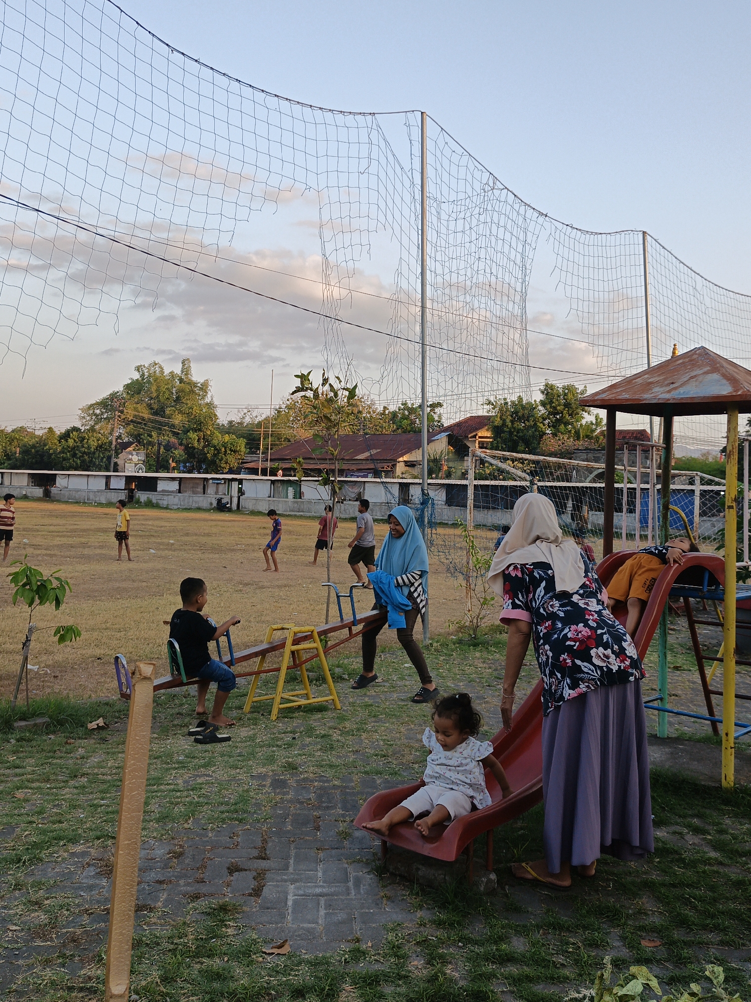 In the city of Trucuk, Indonesia, a playground is depicted with a tattered volleyball net overhead. In the foreground, a young girl sits on a red slide, accompanied by two women. To the left, a boy sits on a seesaw, while other children are visible in the background playing soccer on a field. Trees and buildings line the perimeter of the field, under a sky with scattered clouds.