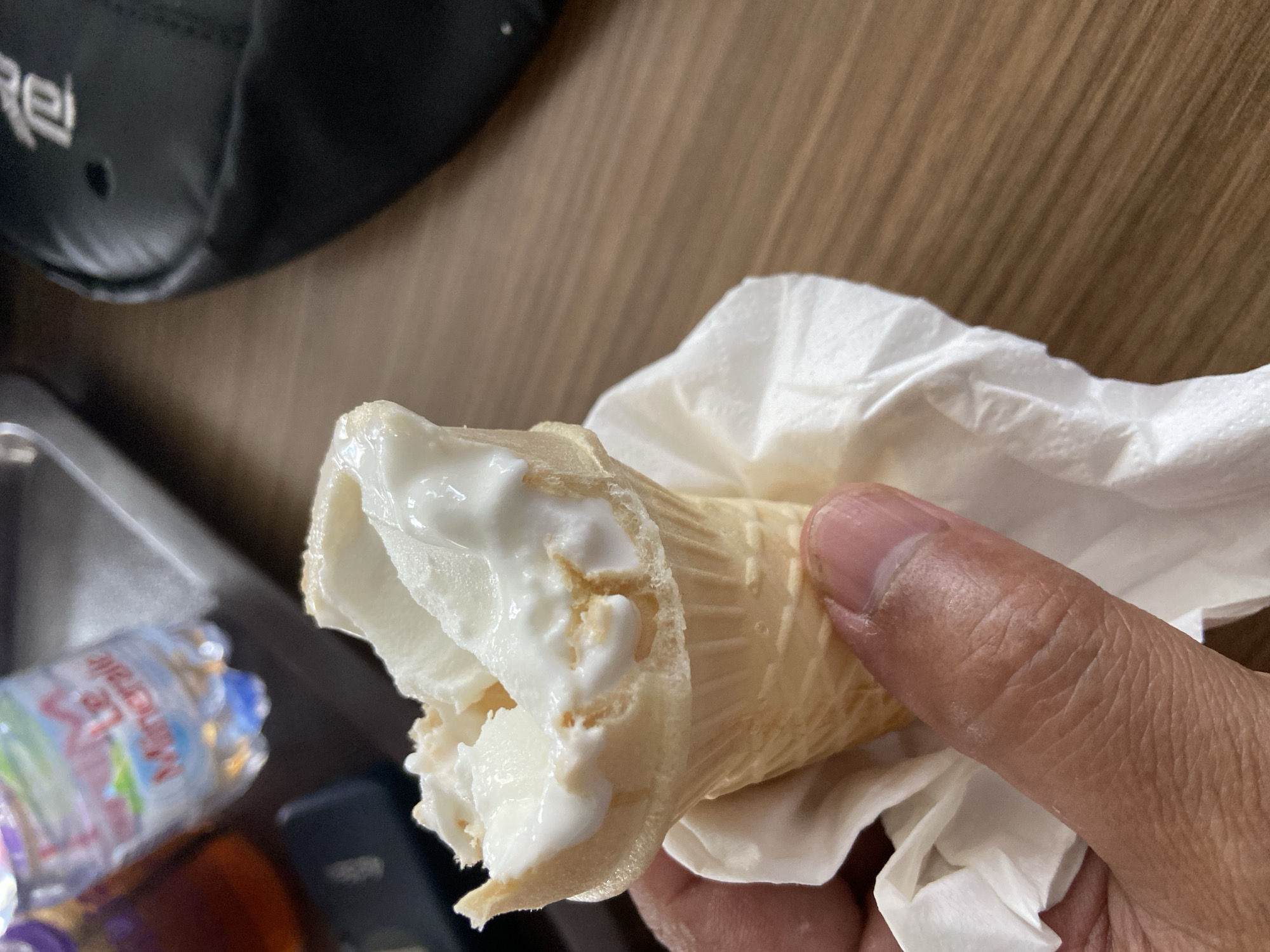 In Bago, Indonesia, a close-up shot focuses on a person's hand holding a partially eaten vanilla ice cream cone, wrapped in a white napkin. The ice cream is melting, with drips cascading down the waffle cone. In the background, a bottle of Evian mineral water and a black bag are visible, resting on a wooden surface. The scene suggests a casual setting, possibly a cafe or a picnic.