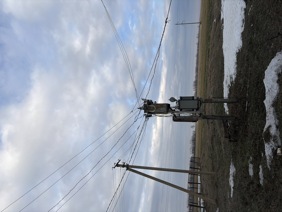An outdoor scene depicts an electrical transformer mounted on poles, with power lines extending across a cloudy sky. A field with remnants of snow is adjacent to the infrastructure. A utility pole with angled support is visible, along with a fence line. The horizon is punctuated by distant trees.