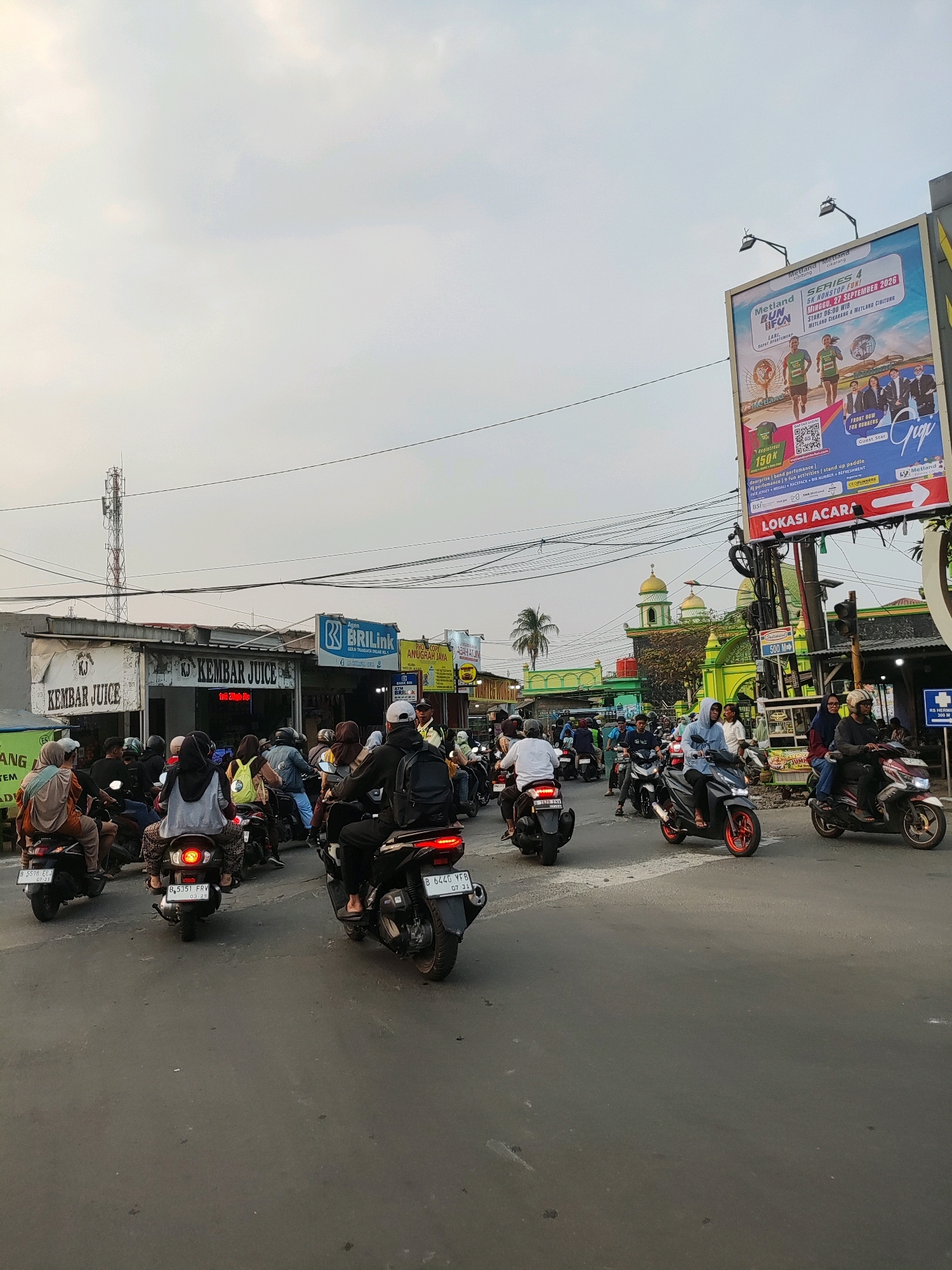 In Telagaasih, Indonesia, a traffic jam with numerous motorcycles is captured on a street. The scene is dominated by a dense formation of motorcycles, many with riders wearing helmets and casual attire, moving in the same direction. The road surface is asphalt, and the atmosphere suggests a warm, possibly humid, climate given the clear sky with some scattered clouds.
On the left side of the road, a building with a sign reading "KEMBAR JUICE" is visible, indicating a commercial establishment. Further down, a BRI Link branch is advertised. Across the street, a bright green mosque with golden domes stands prominently, a significant landmark in the area. A large billboard advertising a "METLAND RUN & FUN SERIES 4" event is positioned above the street on the right, featuring images of runners and performers. Traffic lights and utility poles with wires crisscross the sky. The overall impression is one of a bustling urban environment with heavy motorcycle traffic, typical of many Indonesian cities.