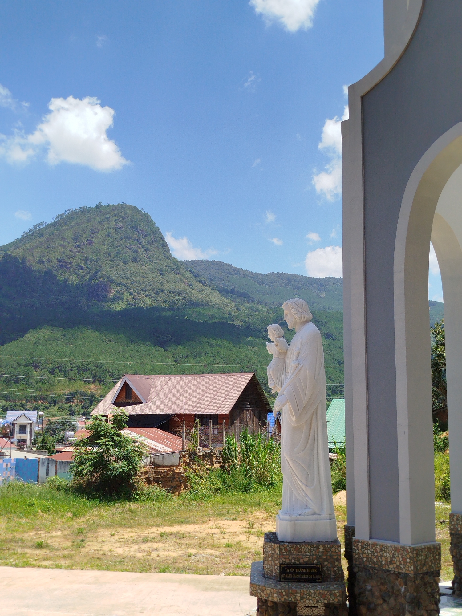 In K'Rèn, Vietnam, a white marble statue of Jesus holding a child stands to the right of the frame, partially obscured by a white archway. The statue is on a pedestal with a plaque that reads "TA ON THANH GIUSE / GD MARIA HOANG THI KIM CHI DA LAT". Behind the statue, a lush green mountain range dominates the background under a bright blue sky with scattered white clouds. In the middle ground, there are several buildings, including a house with a rusty brown corrugated metal roof and other residential structures. The foreground consists of a dirt and grass area.