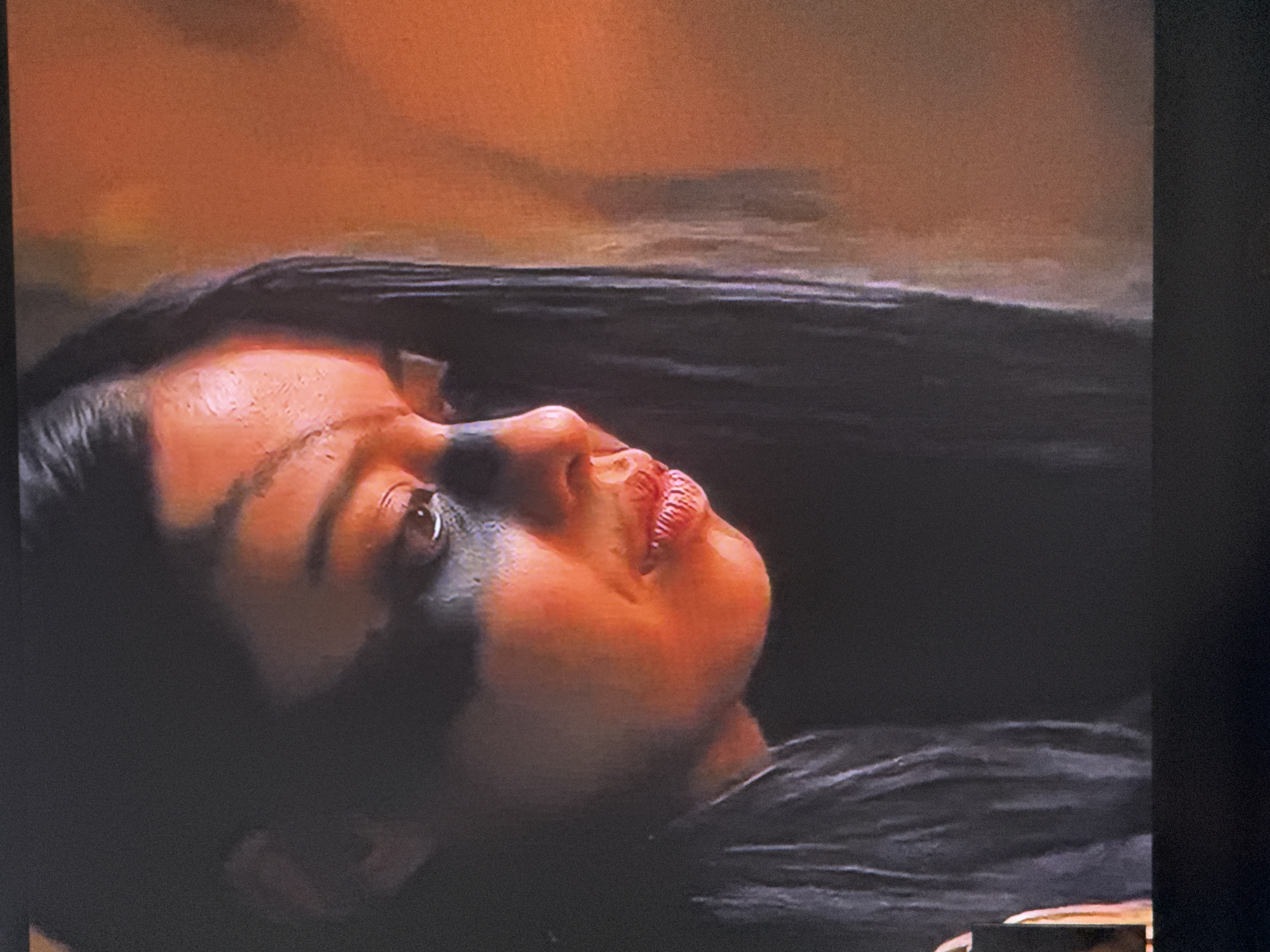 The image shows a close-up of a woman's face, angled upwards. She has dark hair, fair skin with a reddish tint, and black markings under and around her left eye. Her lips are red. Her gaze is directed to the upper right of the frame. The background behind her is blurry and appears to be a mottled orange and brown color. The lower right portion of the frame shows a dark textured surface which might be her clothing. No location in Tbilisi, Georgia is evident.