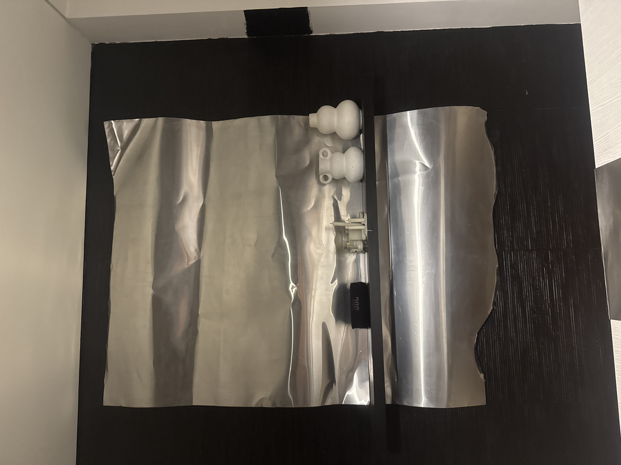 The image shows a dark, textured wall with two large sheets of shiny, silver material hung on it. One sheet is larger and covers most of the left side of the wall, while the other is narrower and positioned on the right. Between the two sheets, near the top and center, are three white, plastic objects that appear to be plumbing or fixture components. Further down, attached to the silver material, is a small black object with the white text "JBL" visible. The overall impression is of an art installation or display, possibly in an indoor setting. There are no people or obvious indications of weather or time of day. The location context of Kuta Selatan, Indonesia, does not provide any specific visual cues within the image itself.