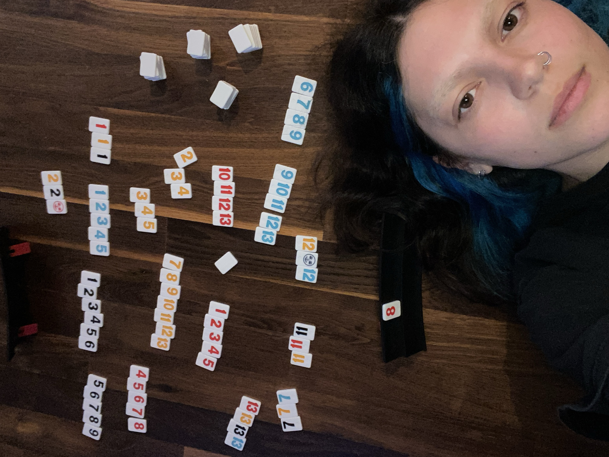 A woman with blue-tinged dark hair lies on a dark wooden floor beside Rummikub tiles arranged in runs and groups. A black tile rack with an "8" tile sits near her shoulder. The tiles include runs of 1-13 in various colors, groups of the same number, and some loose tiles. The floor's surface appears to be a dark, stained wood, and the room is dimly lit. The woman is looking at the camera with a neutral expression and has a nose ring. There is no information in the image to suggest that the location is in the city of Villaviciosa, Spain.