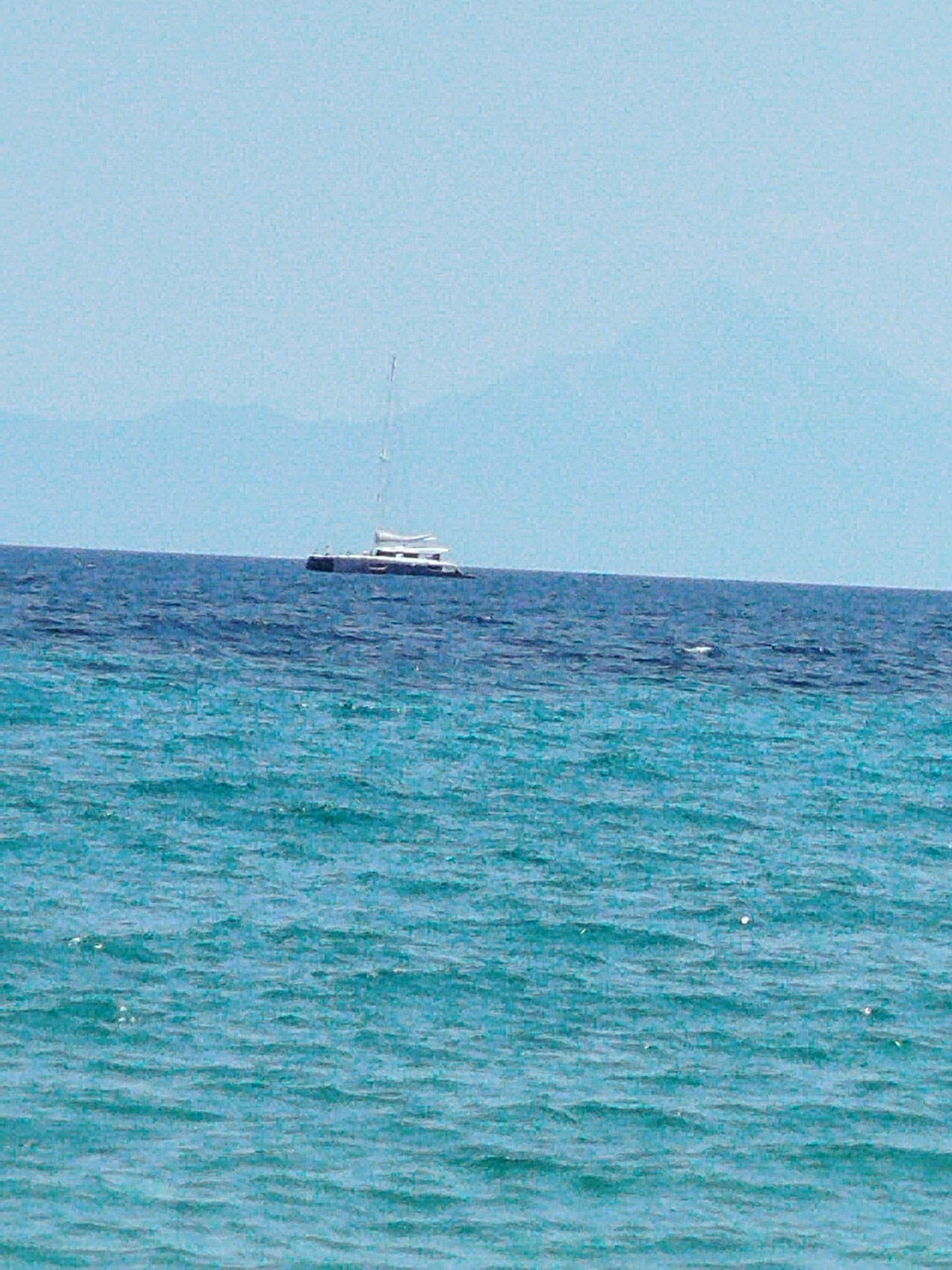 A wide shot captures a vast expanse of the Aegean Sea under a bright, pale blue sky. In the distance, hazy mountain ranges form a faint horizon line. A white catamaran with a single mast is positioned on the darker blue water, appearing as a solitary object against the sea and sky. The foreground is dominated by the vibrant turquoise waters of the sea, with gentle ripples and waves. The scene appears to be located off the coast of Sithonia, Greece, given the characteristic clear blue waters and distant mountainous landscape.