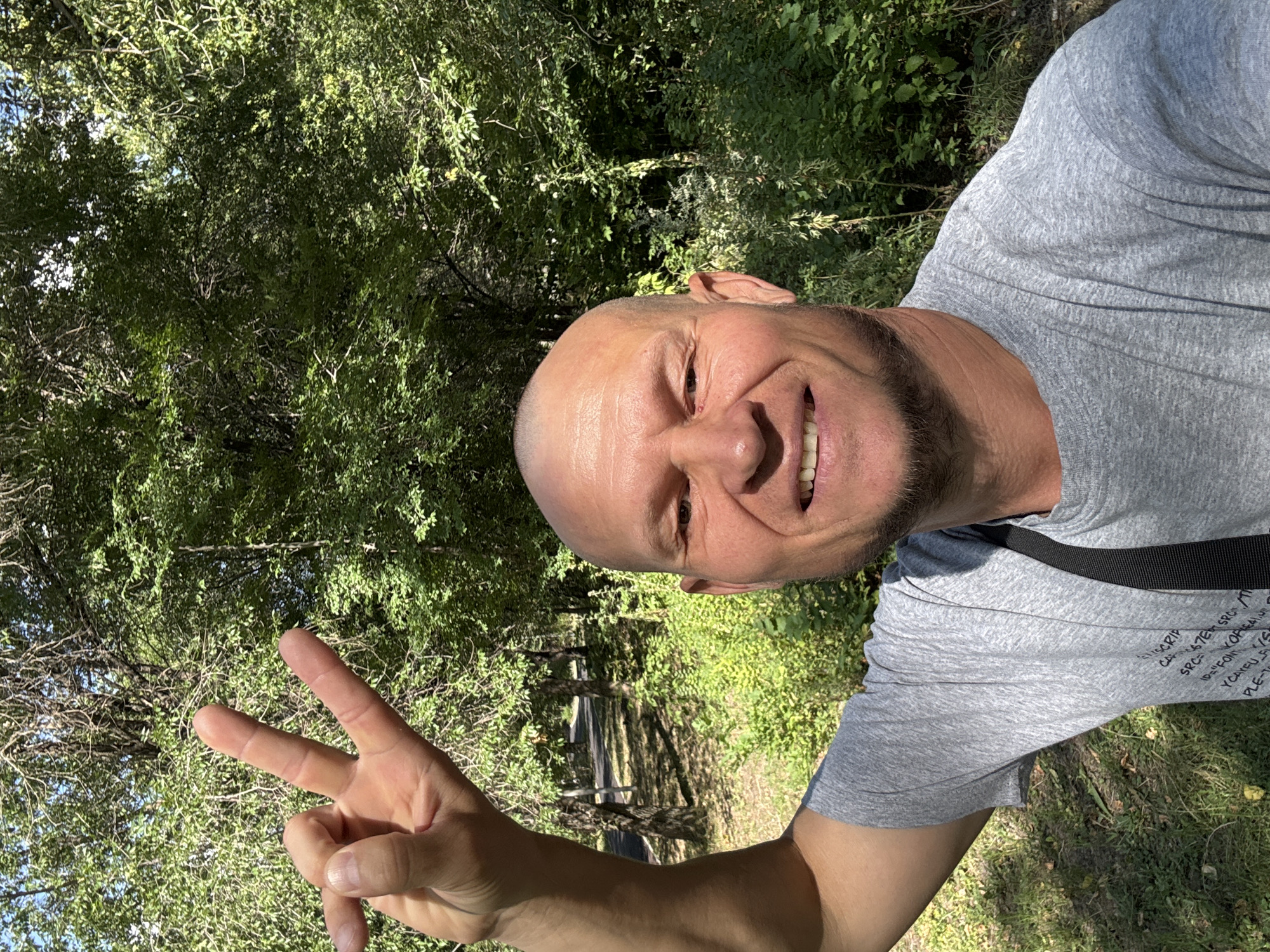 A man with a shaved head and a goatee is taking a selfie outdoors. He is wearing a grey t-shirt and has his left arm raised, displaying a peace sign with his fingers. He is smiling broadly and looking at the camera. The background consists of lush green trees and foliage, suggesting a park or wooded area. Sunlight filters through the leaves, creating dappled shadows on the ground. The man's t-shirt has some text printed on it, but it is mostly obscured. The scene appears to be set during the daytime. The exact location within Oskemen, Kazakhstan, cannot be determined from the provided image.